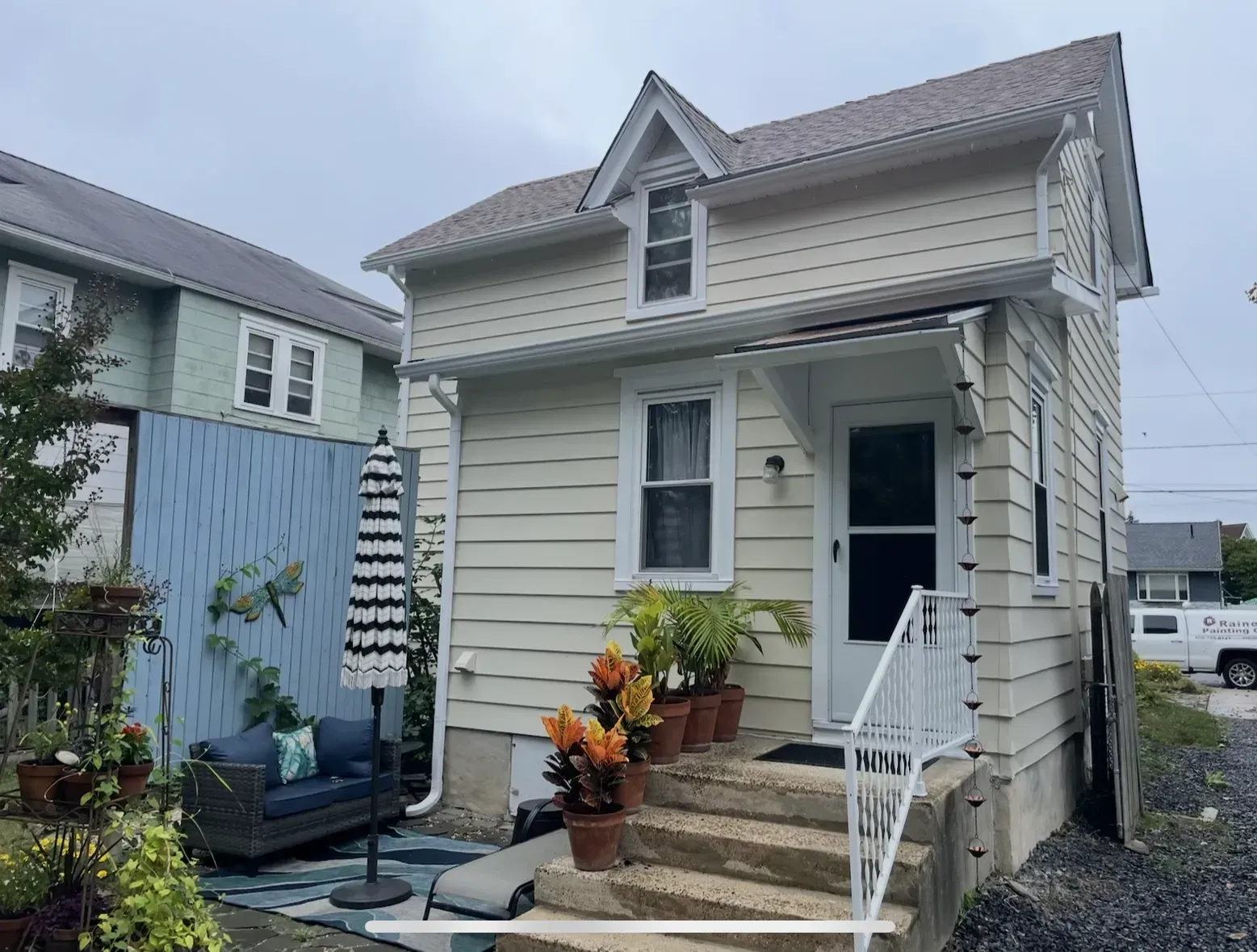 Beige house with a small porch, steps, and potted plants. A blue fence and outdoor seating are visible.