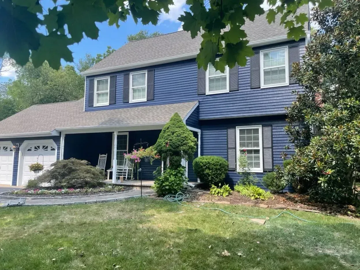 Two-story blue house with white trim, shutters, and garage doors; front yard with greenery and blue sky.