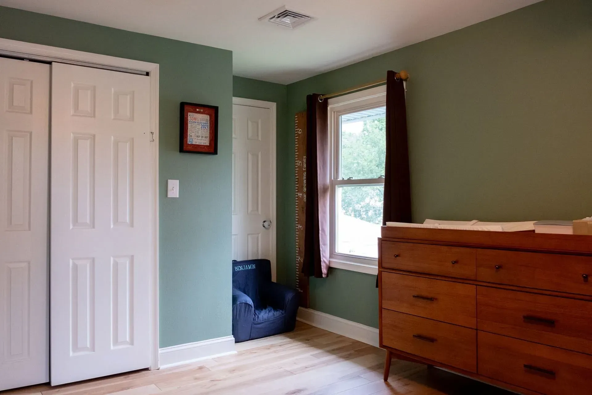 A bedroom with sage green walls, white closet doors, a wooden dresser, and a window.