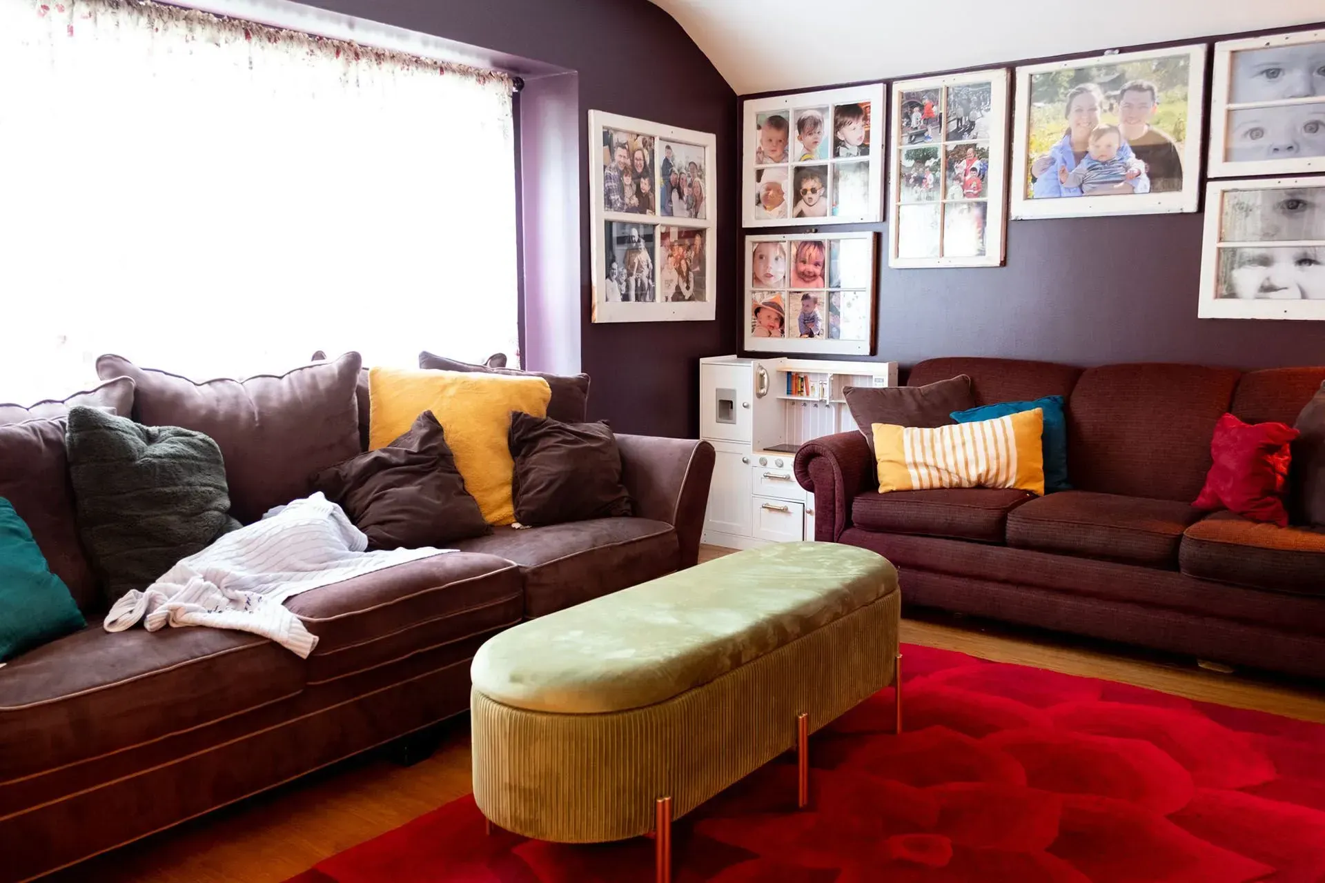Cozy living room with dark brown sofas, colorful pillows, framed photos, red rug, and a green ottoman.