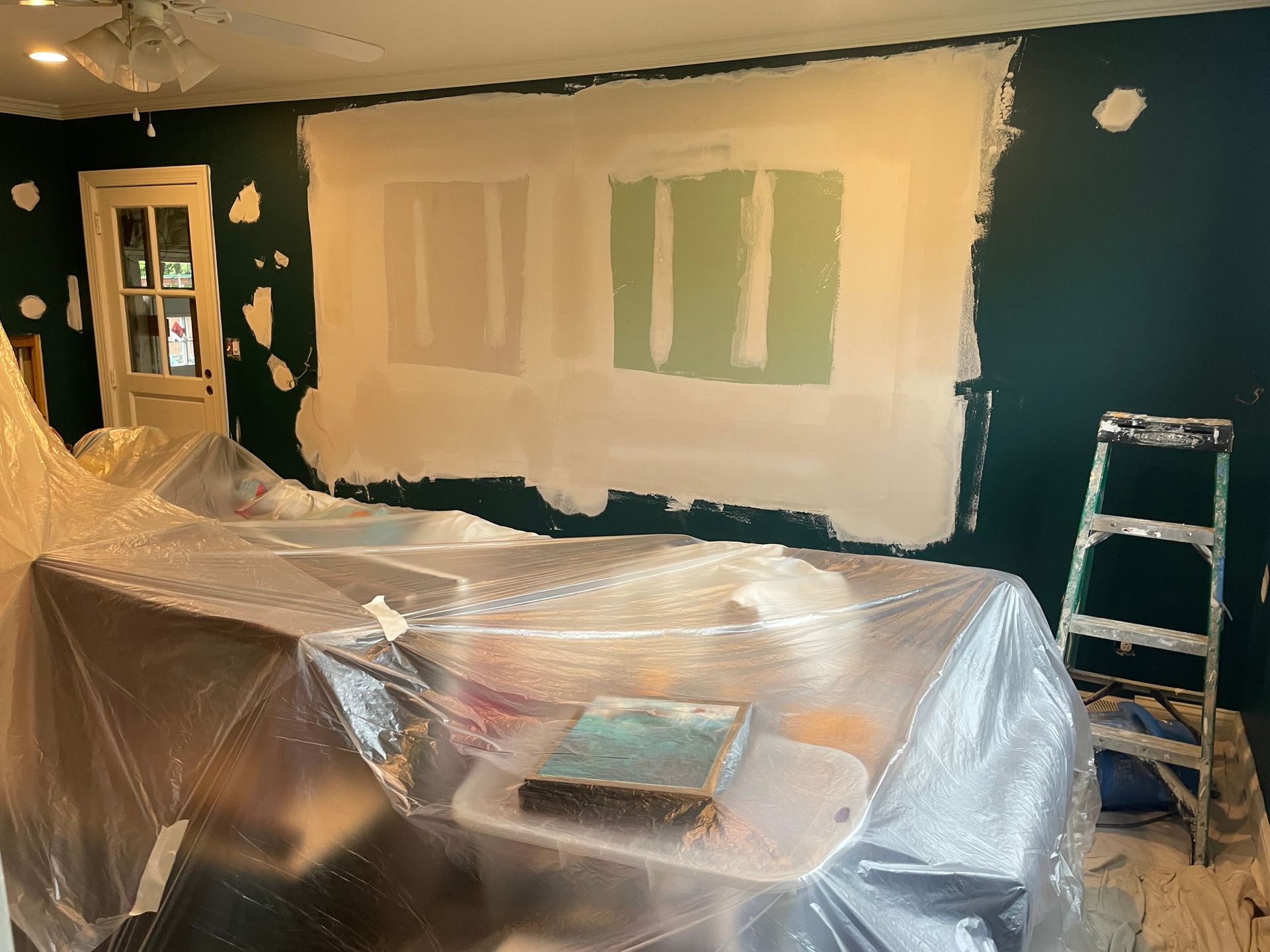 Room interior during renovation; walls with fresh drywall, dark green paint, plastic-covered furniture, and a stepladder.