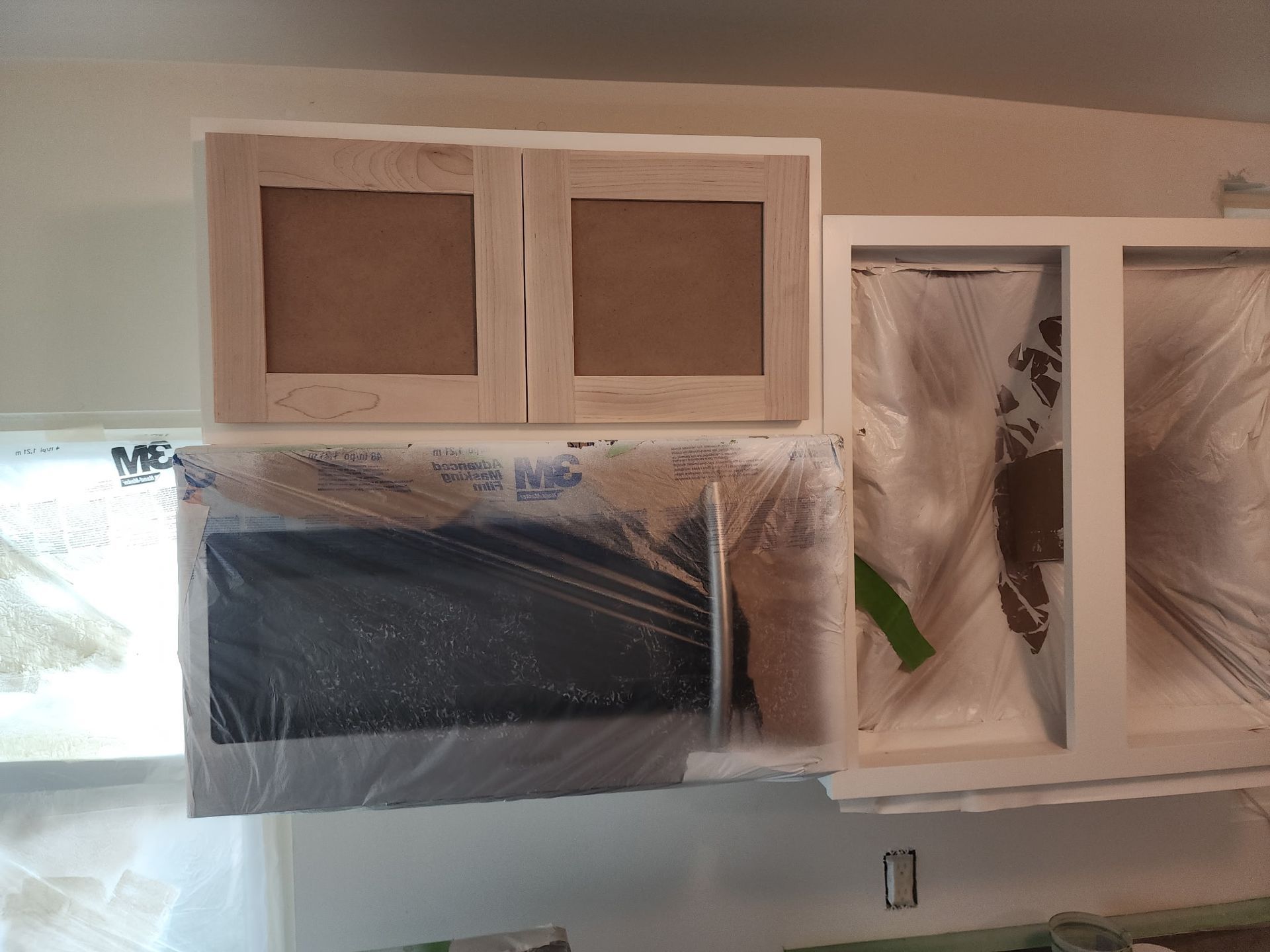Kitchen cabinets being painted; some covered in plastic. Beige walls, wooden cabinet frames, and a microwave are visible.