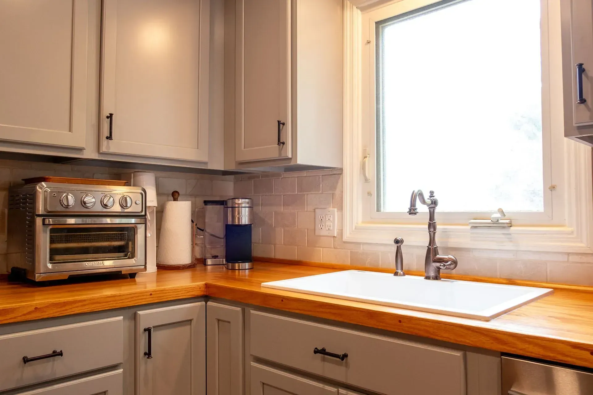 Kitchen corner with gray cabinets, wood countertop, white sink, and window.