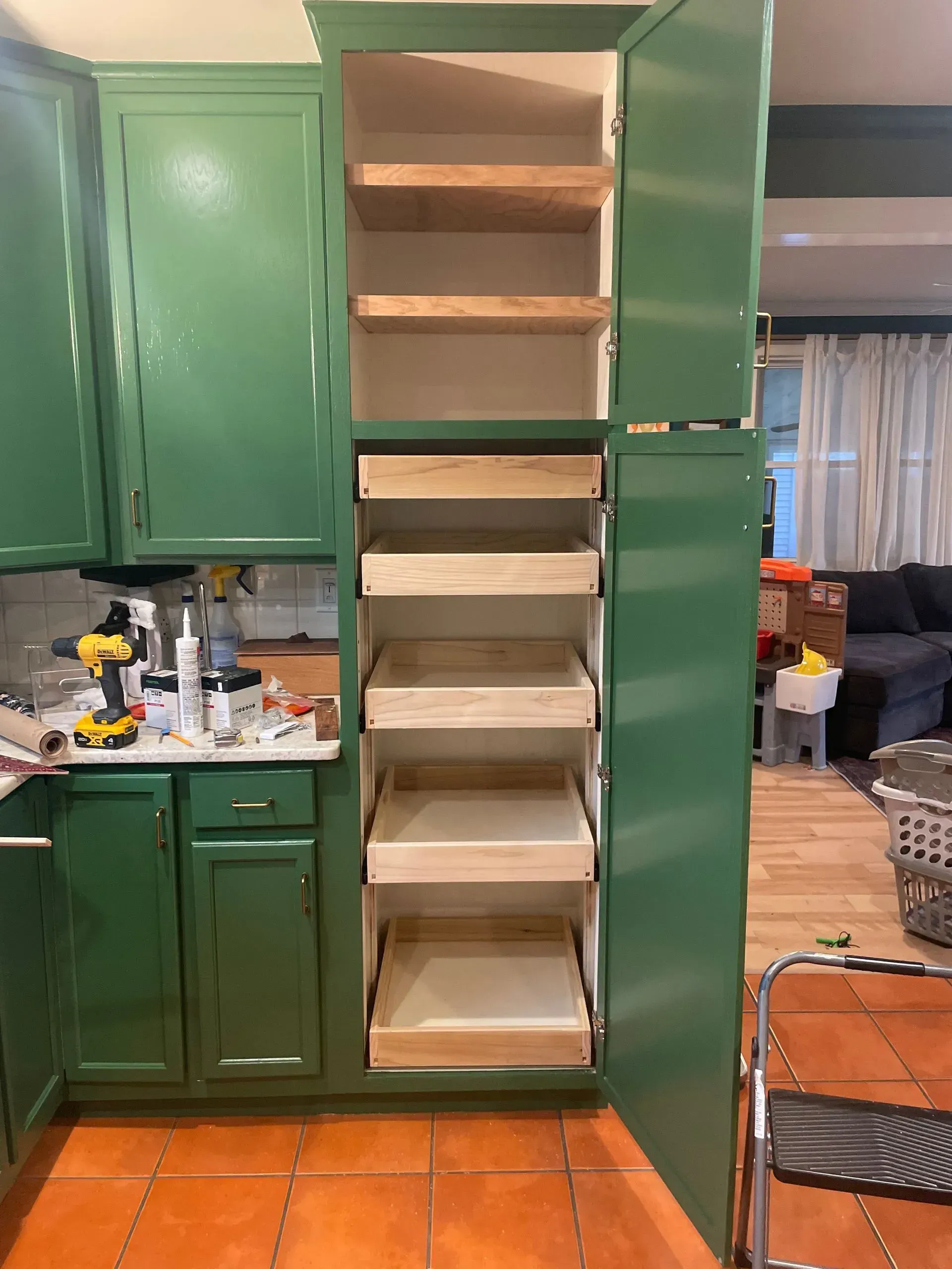 Green kitchen cabinet with open doors revealing pull-out drawers and shelves. A drill and other tools sit on the countertop.