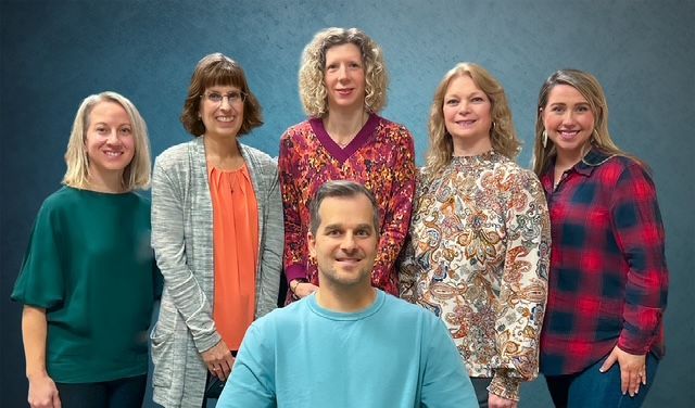 Six people pose in a studio, with five standing behind one seated in front.