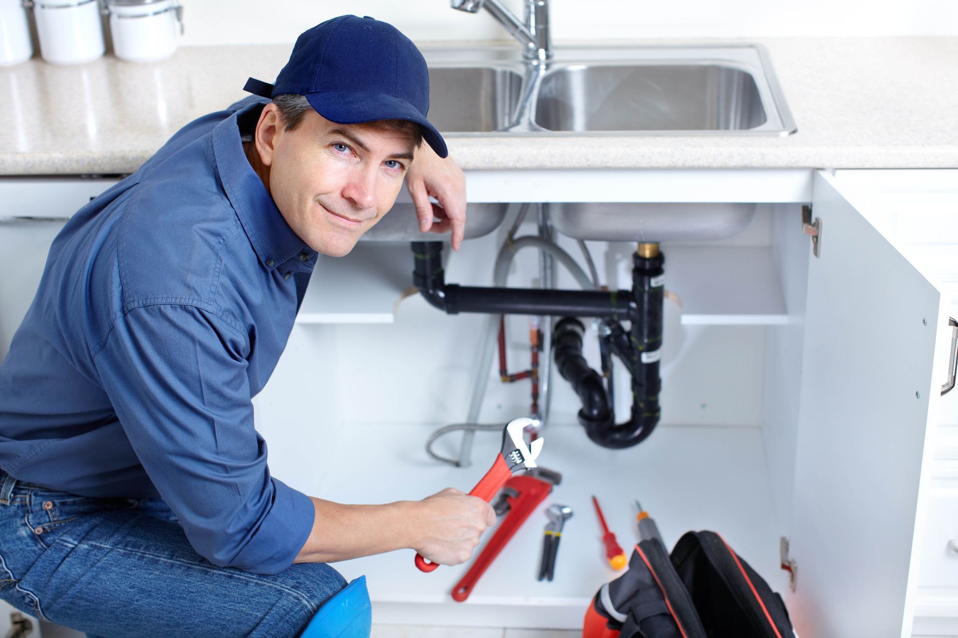 Plumber under a sink, holding a wrench and repairing black pipes in a white kitchen cabinet.