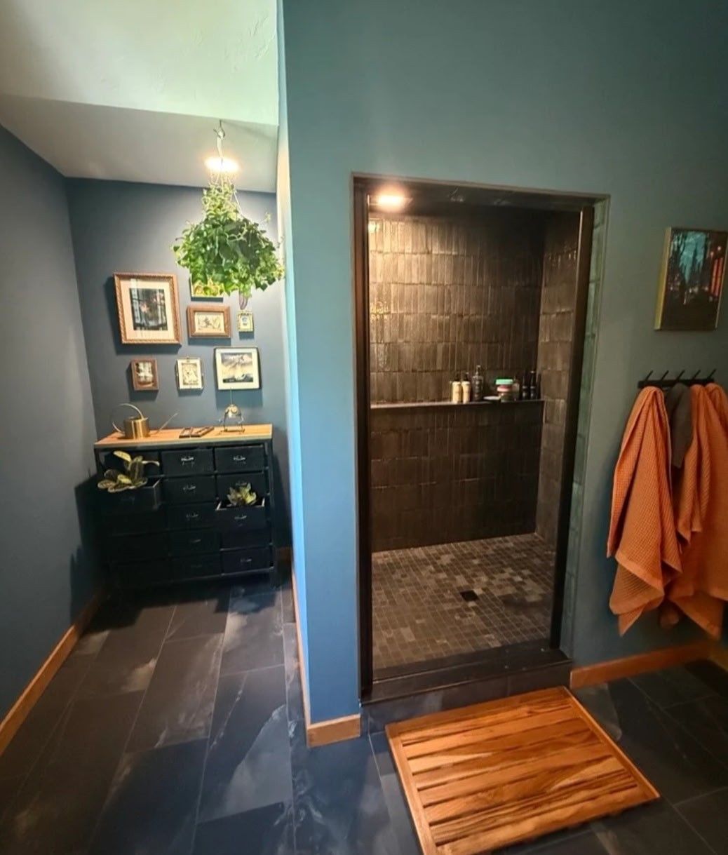 Modern bathroom with dark tiles, a wooden shower mat, and orange towels hanging beside the shower.
