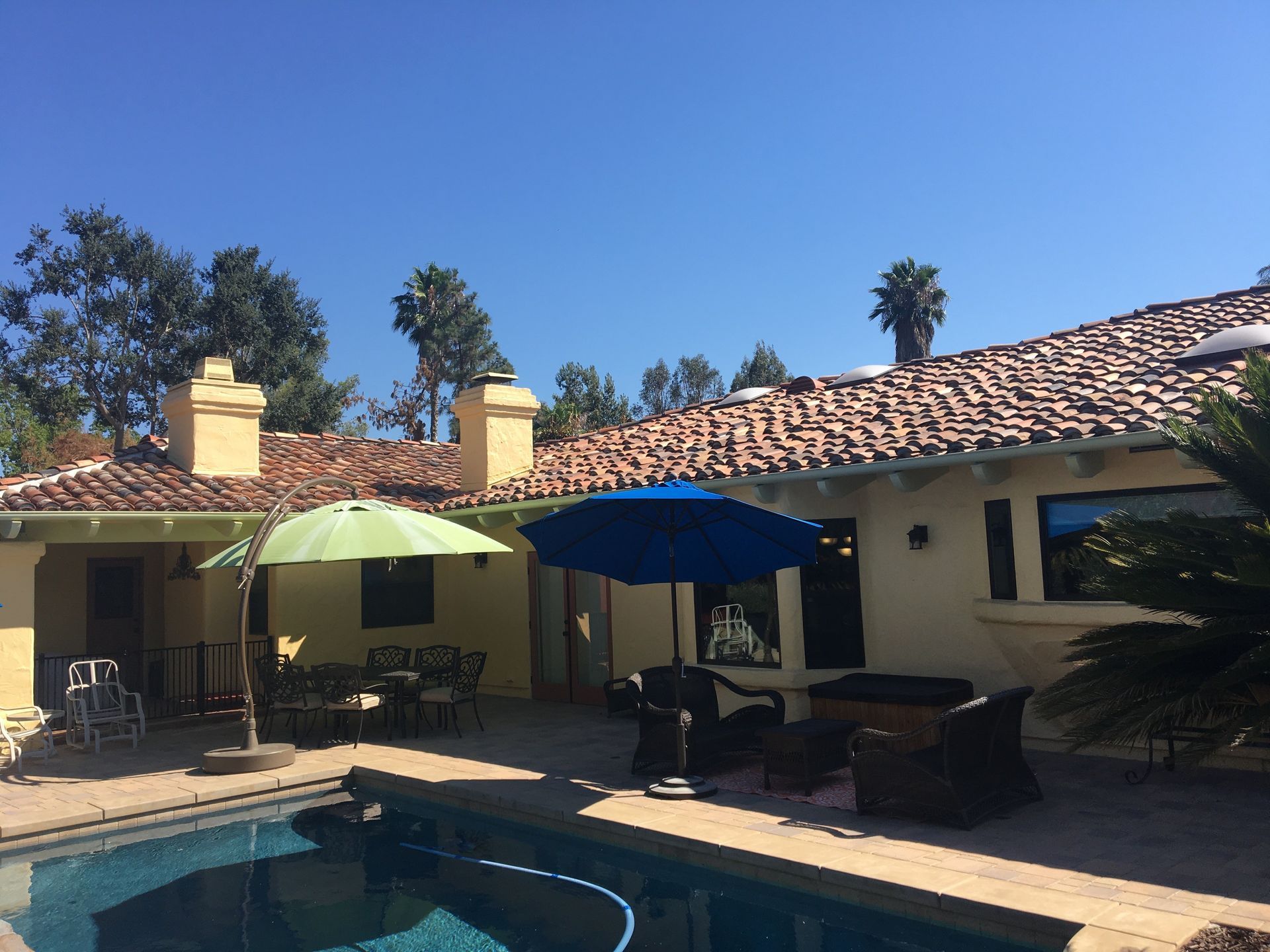 Poolside patio with blue and green umbrellas, beige house, and blue sky.