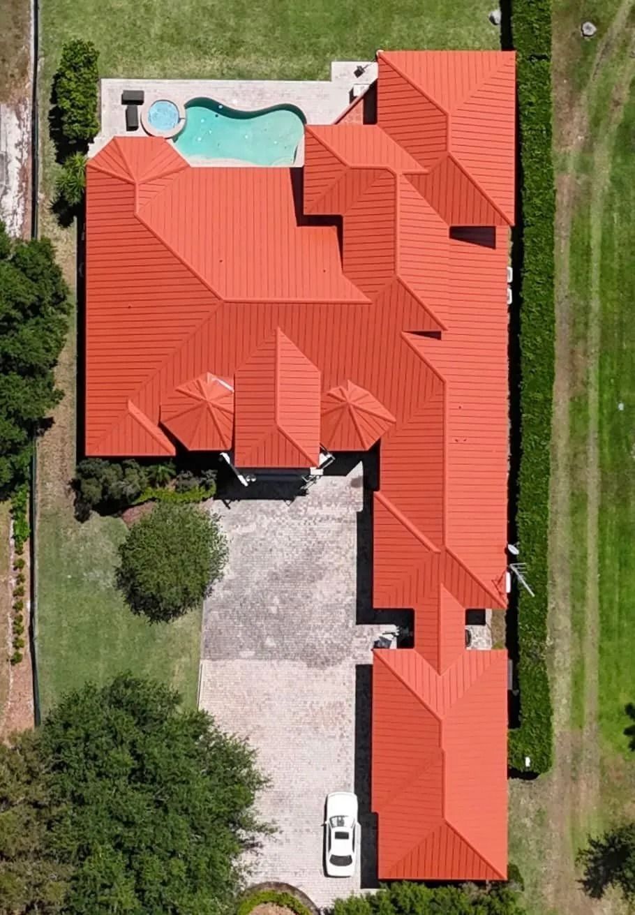 Aerial view of a house with a red roof, pool, and car parked on a gray paved driveway, surrounded by green grass and trees.