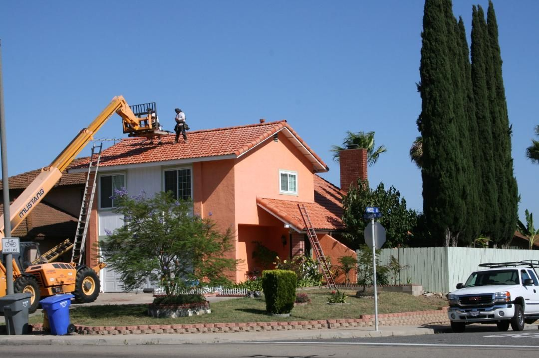 Roofers on a two-story house with orange tile roof, orange walls, and a yellow crane.