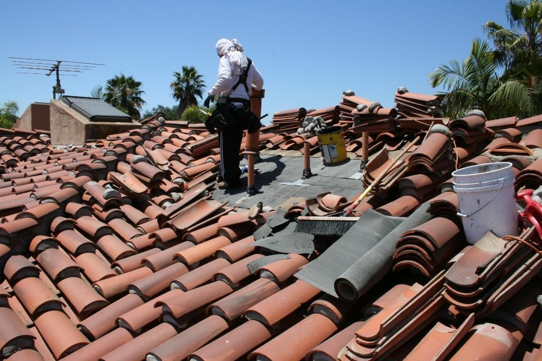 Two people on a tiled roof, removing tiles. Sunny day, terracotta tiles, safety harnesses, and a bucket.