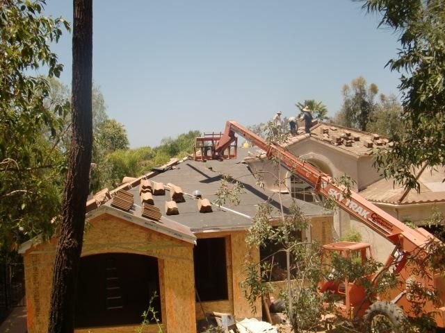 Roofers installing tiles on a residential roof using a forklift, with trees and blue sky.