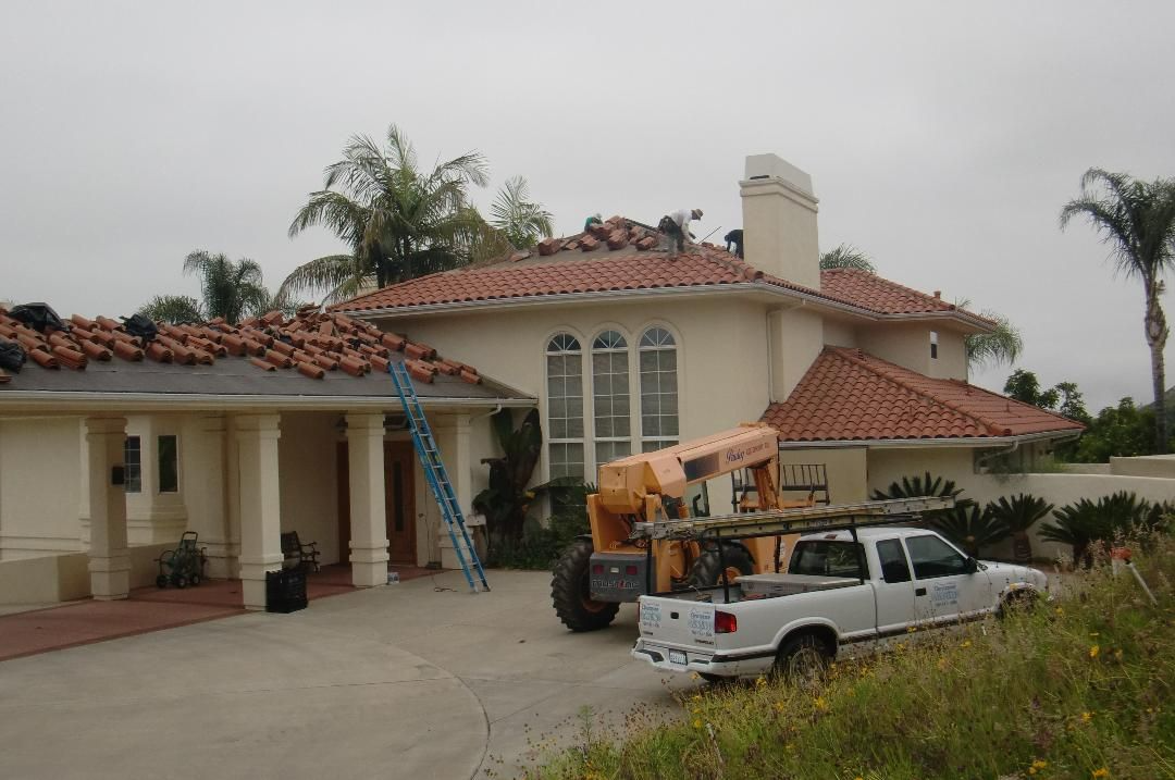 House under construction; workers on roof, orange tiles, white truck and heavy equipment on driveway.