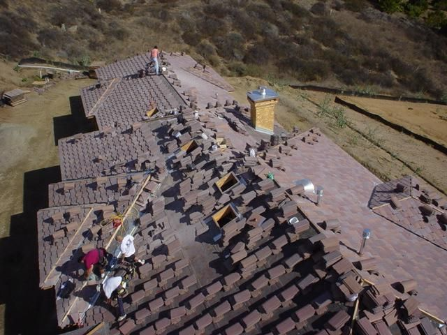 Roofers working on a clay tile roof; brown tiles, tan hill in background.
