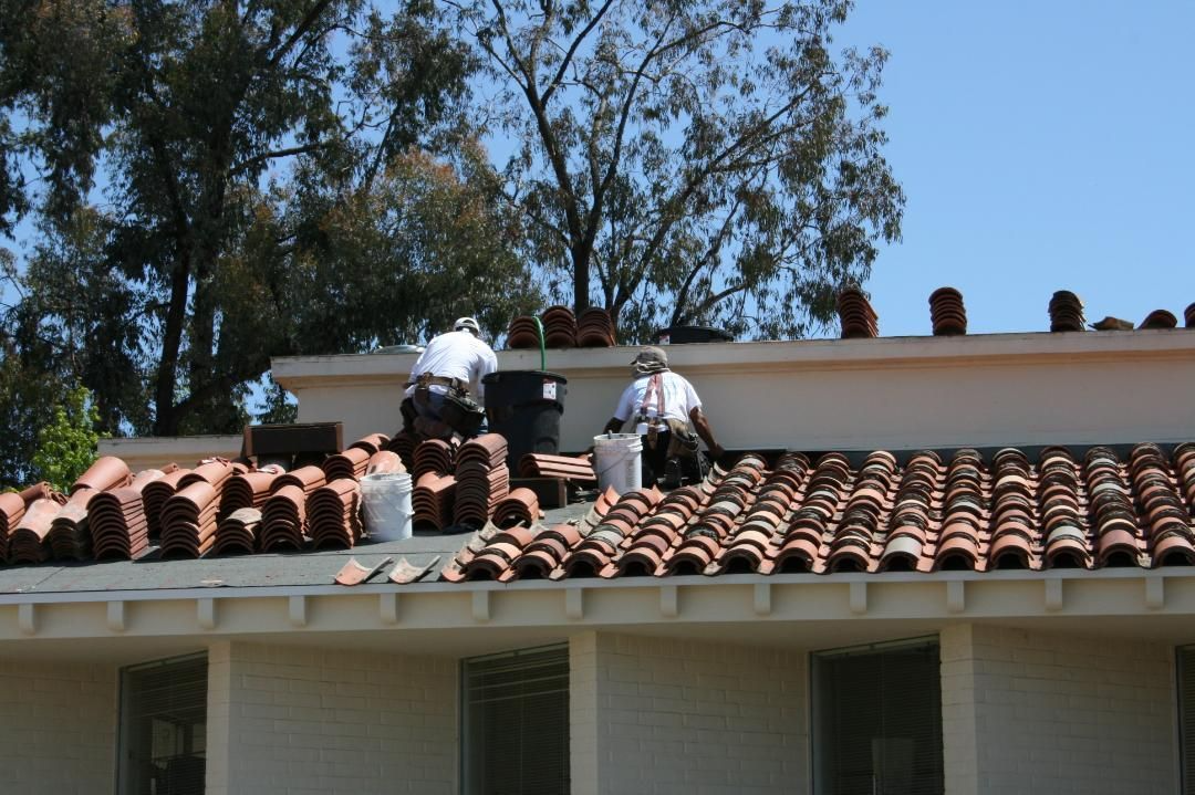 Two roofers replacing clay tiles on a building roof in bright sunlight.