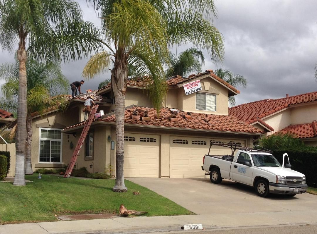 Workers on a tan house with red-tile roof, palm trees, and a white truck in driveway.