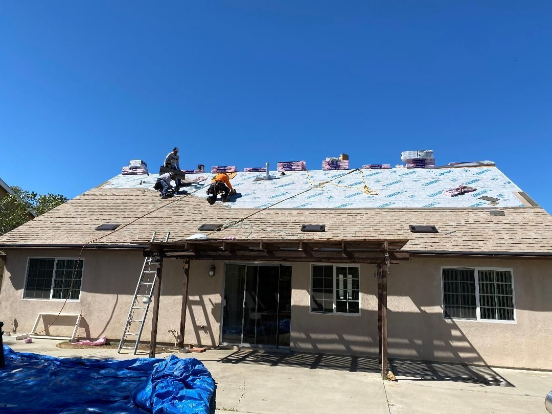 Roofers on a house replacing shingles. Tan house, blue tarp, clear sky.