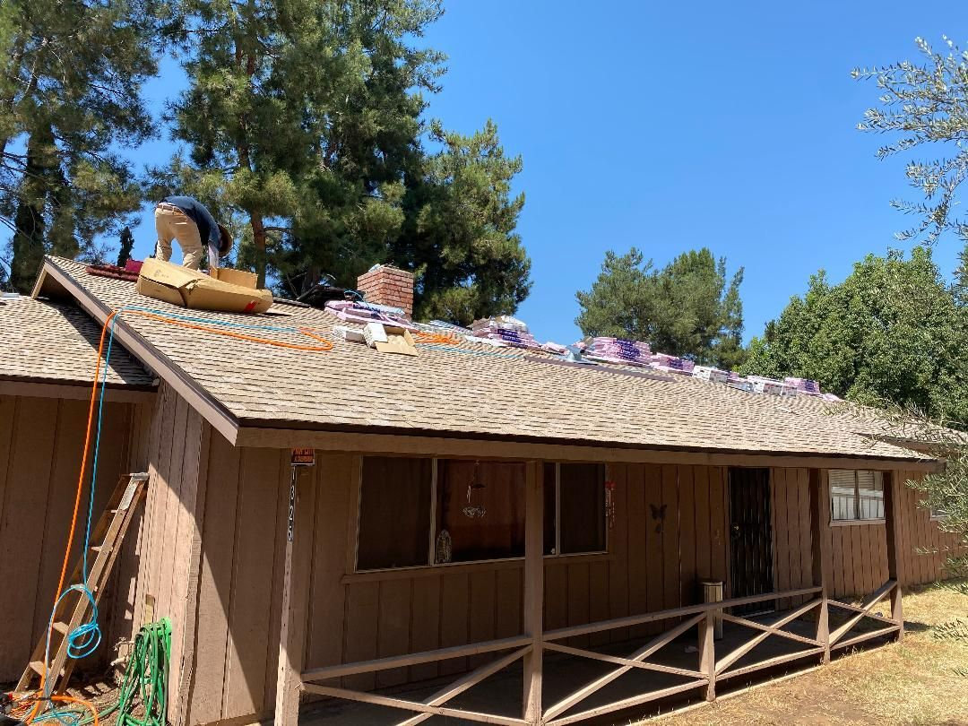 Roofing work on a brown house; a worker on the roof amid shingles and supplies on a sunny day.