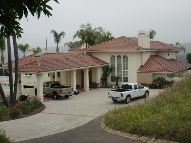 Large beige house with a red-tiled roof, two pickup trucks in the driveway, and a grassy hillside.