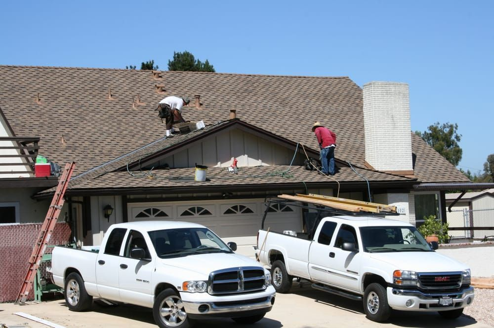 Roofers on a house roof with two white pickup trucks parked in front on a sunny day.