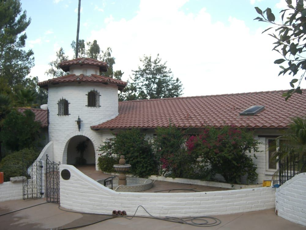 White stucco house with red tile roof and tower, arched entrance, fountain, and gate.