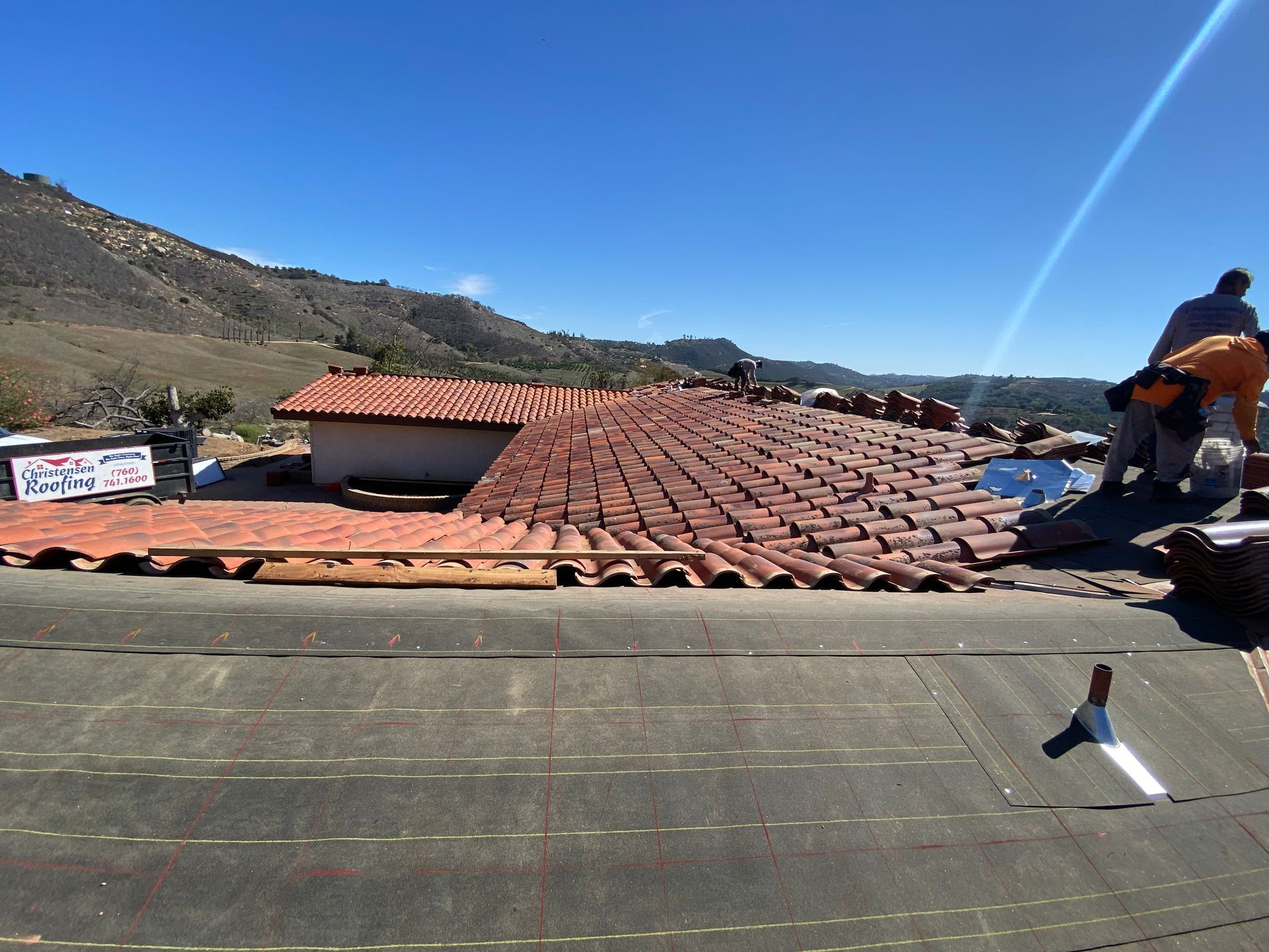 Workers installing terracotta tiles on a rooftop with mountains in the background under a blue sky.