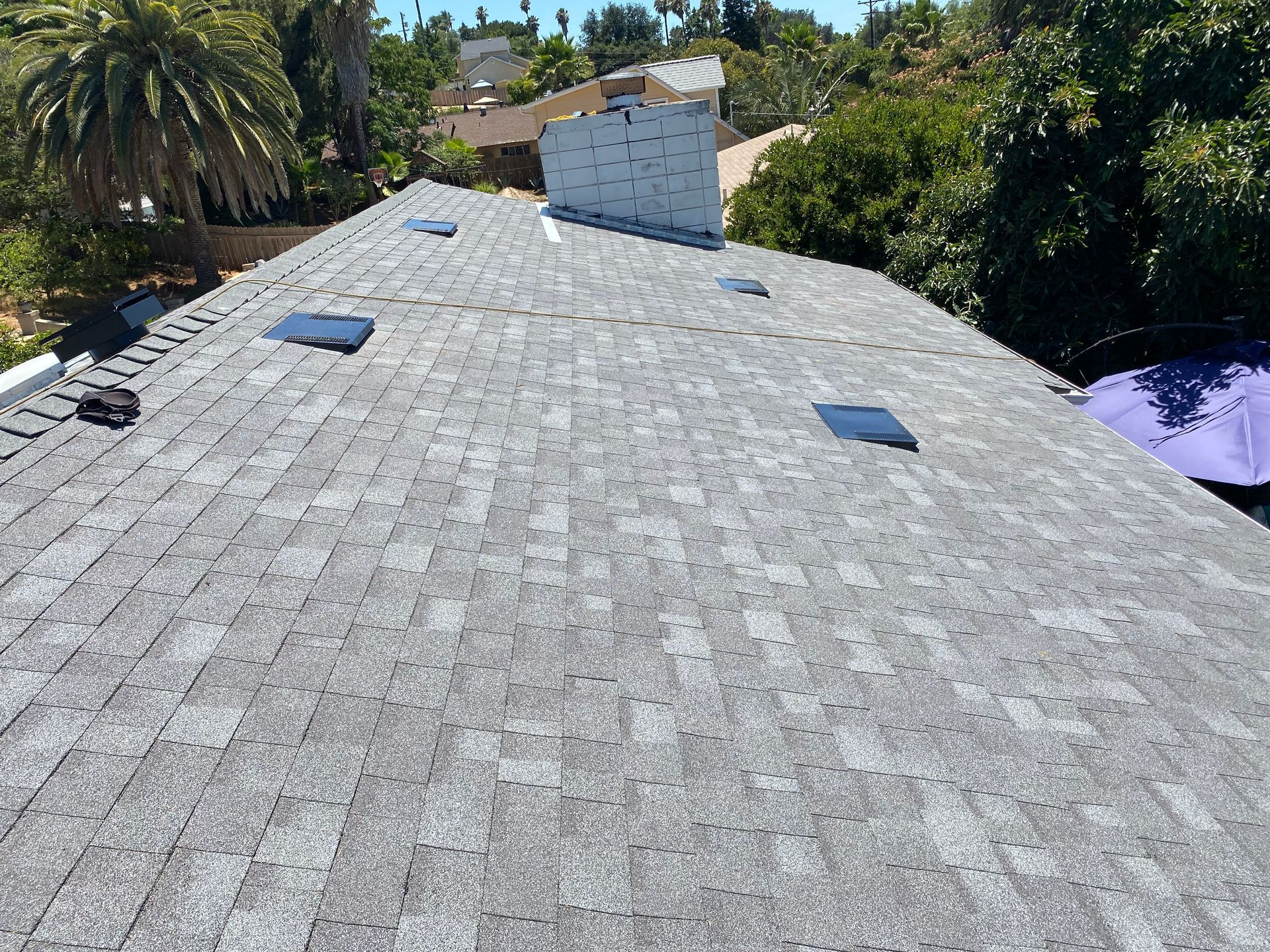 Gray asphalt shingle roof with skylights and chimney, seen from above under a sunny sky.