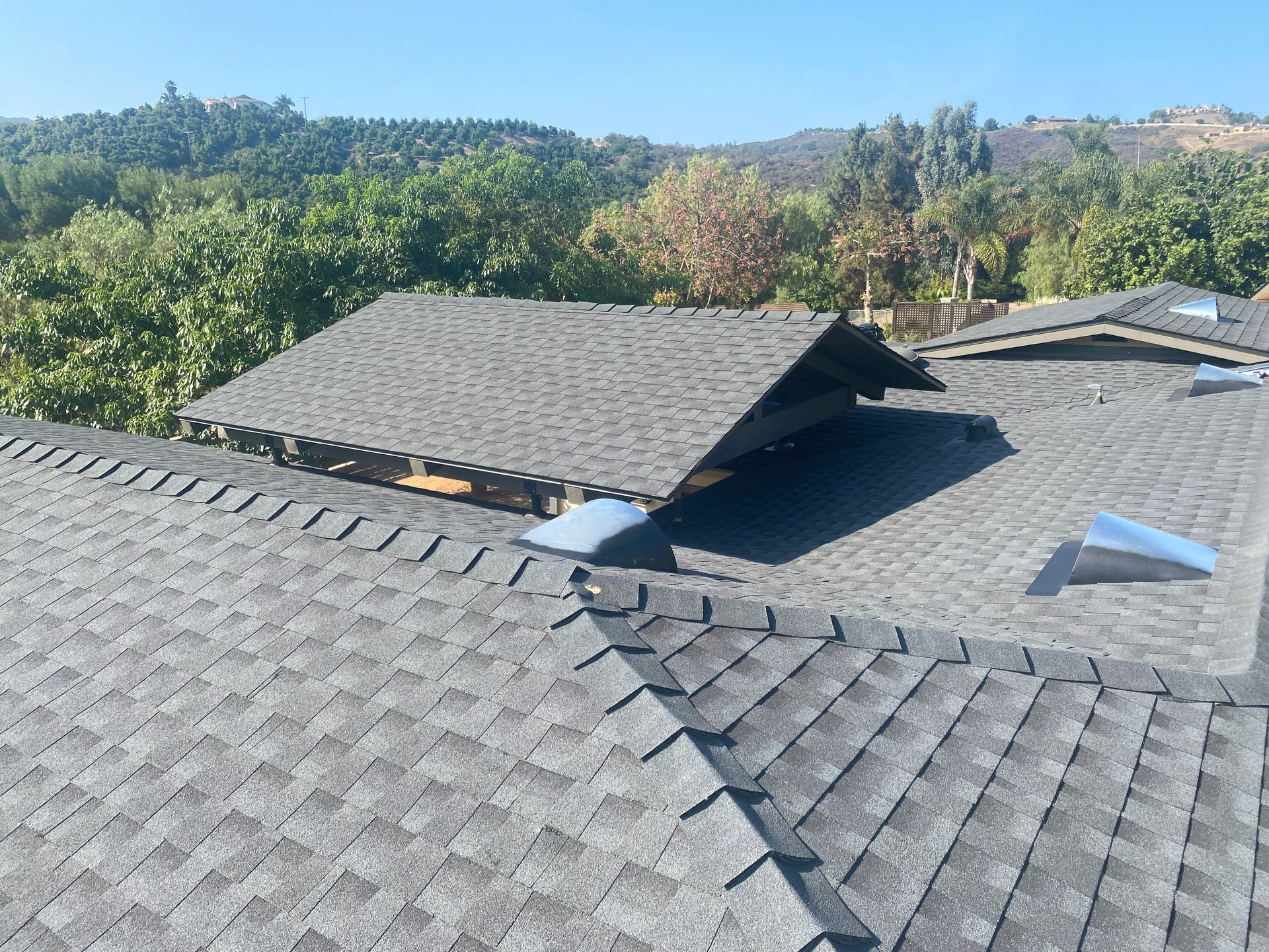 Gray shingled roof with a small gable, and a background of green trees and hills under a blue sky.