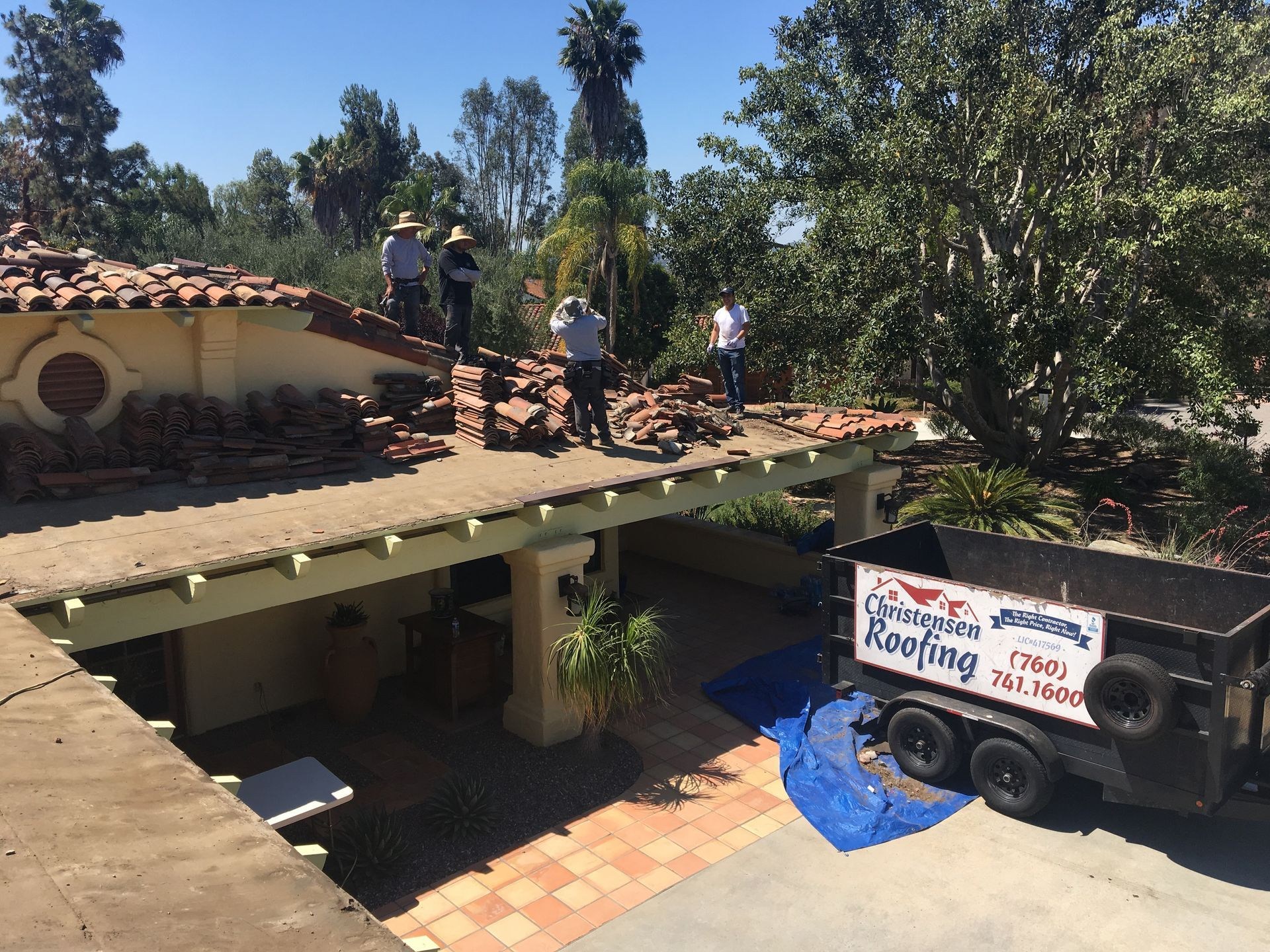 Roofers removing tiles from a Spanish-style house on a sunny day. A trailer for