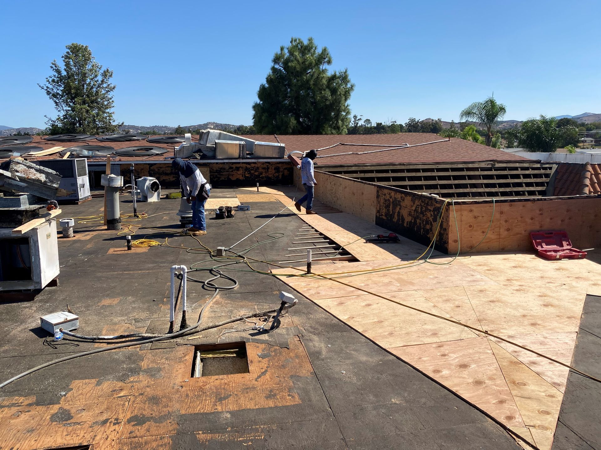 Roofers working on a flat roof, laying plywood and shingles under a sunny sky.