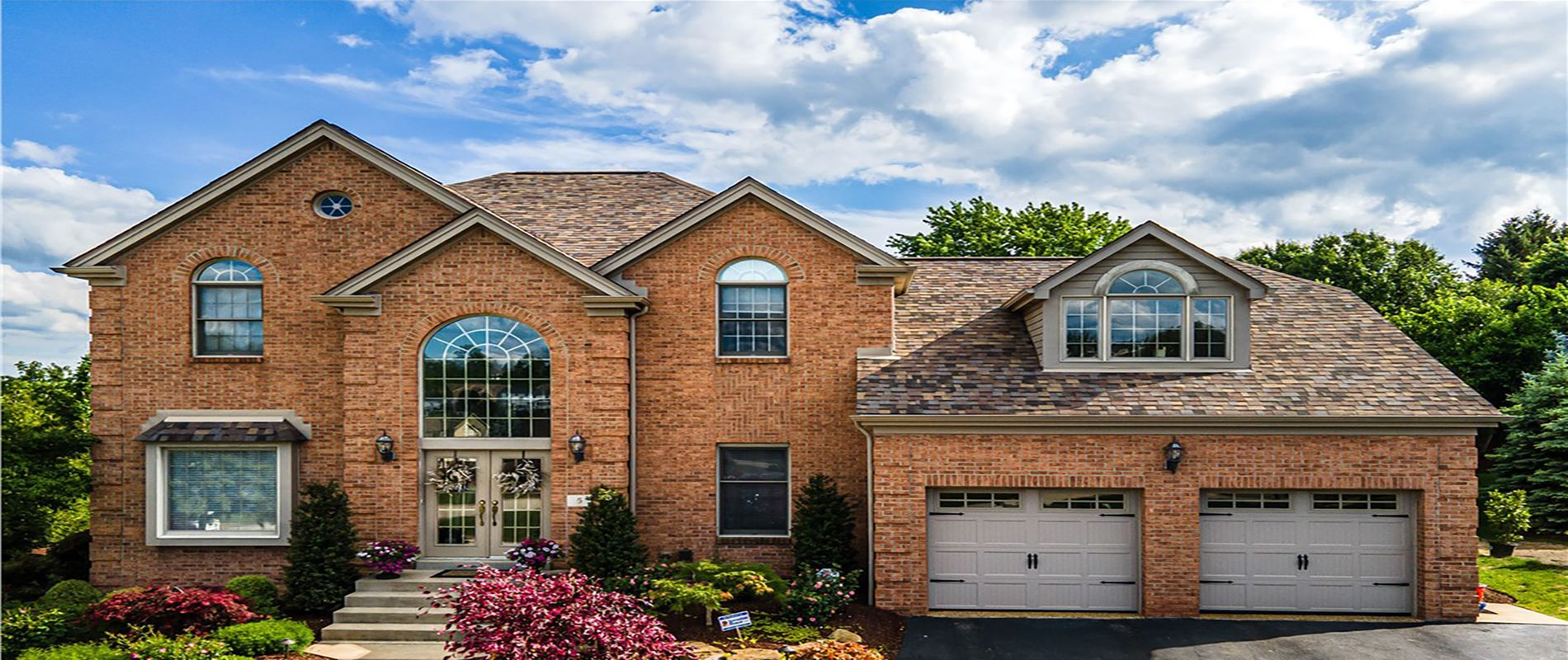 Brick two-story house with arched windows, a two-car garage, and colorful landscaping under a cloudy sky.