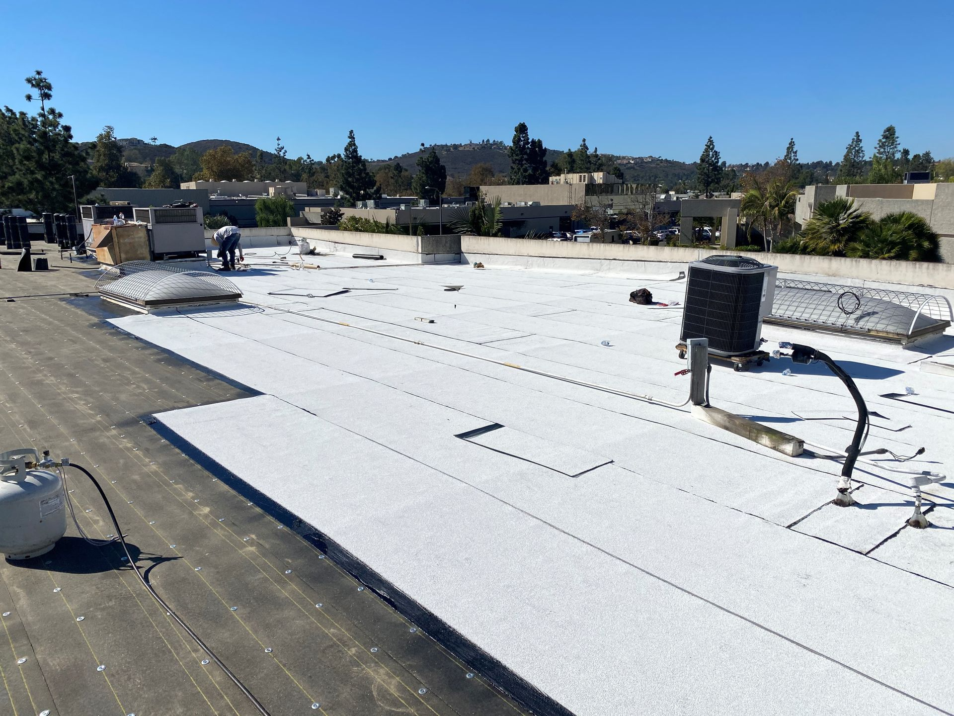 Roof being repaired with white material; propane tank, equipment, and person on roof. Hills and buildings in background.