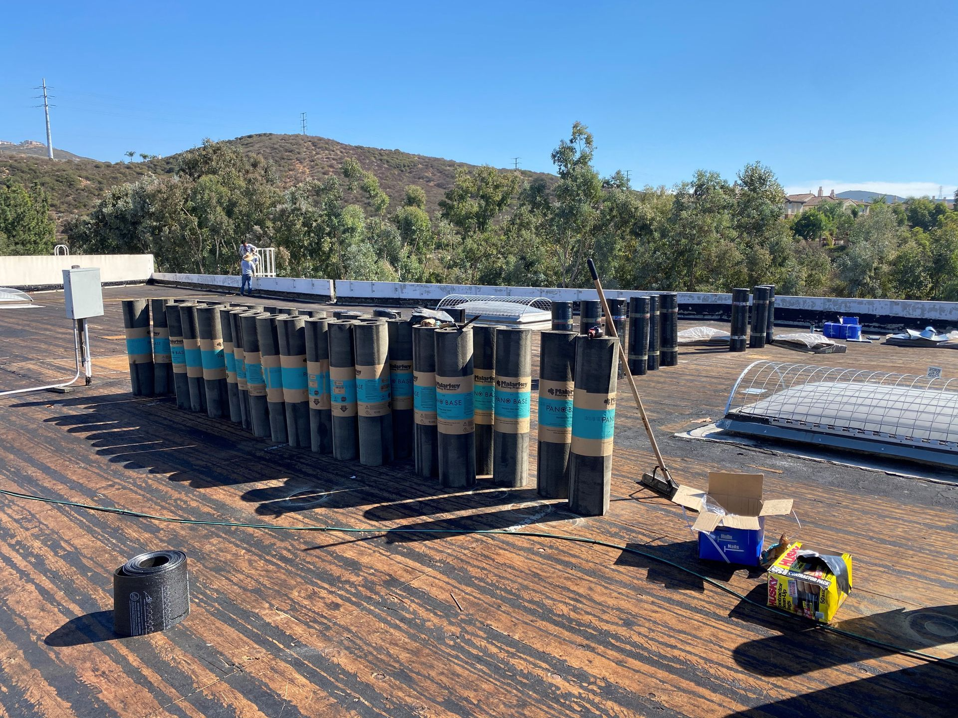 Roofing materials stacked on a flat roof, ready for installation, with a backdrop of trees under a blue sky.