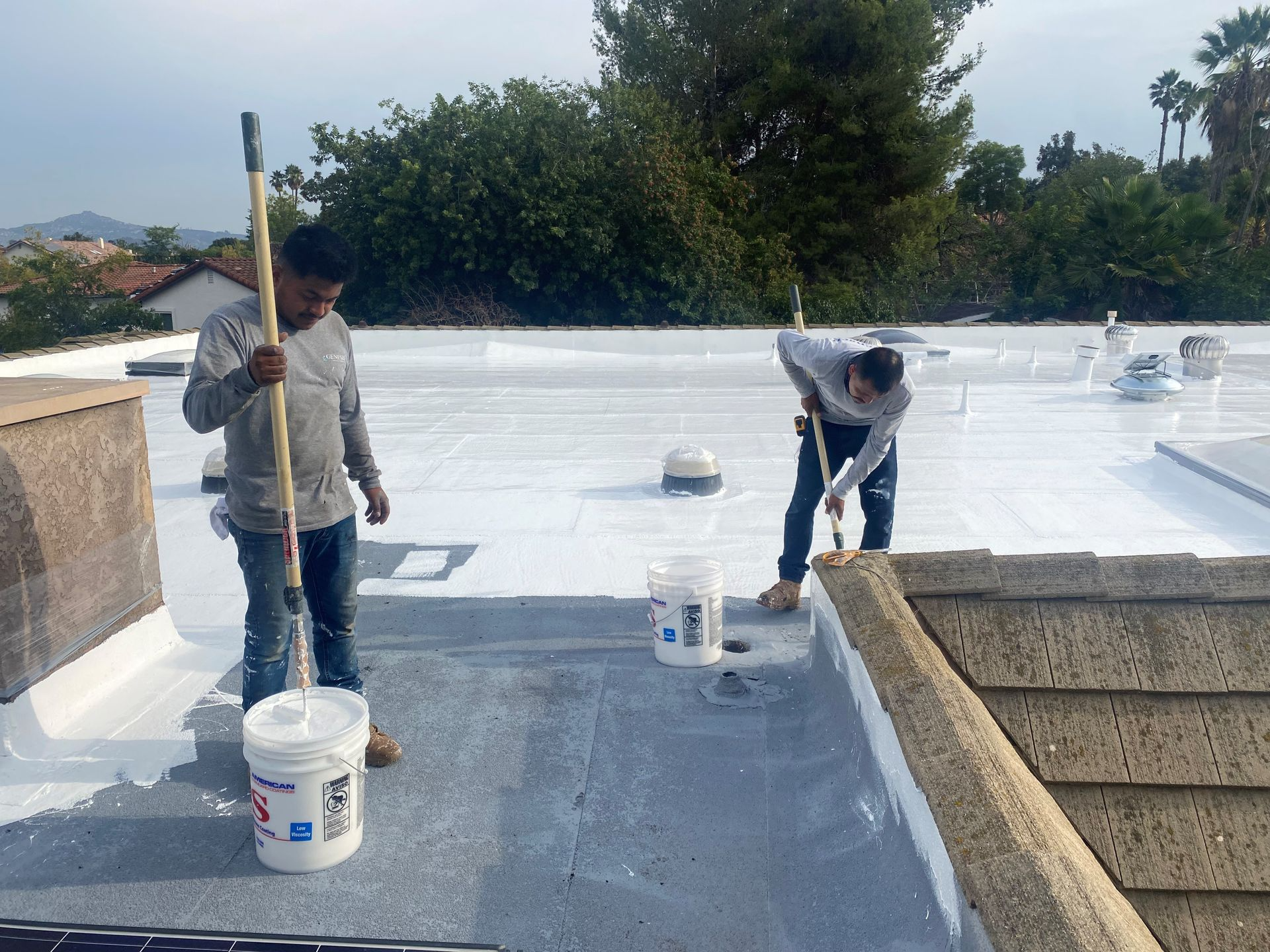 Two workers painting a flat roof white, one using a roller, the other a brush.