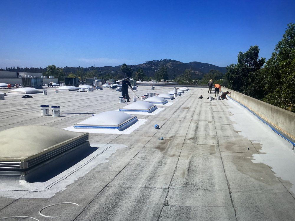 Workers applying sealant to a flat commercial roof with skylights, under a clear blue sky.