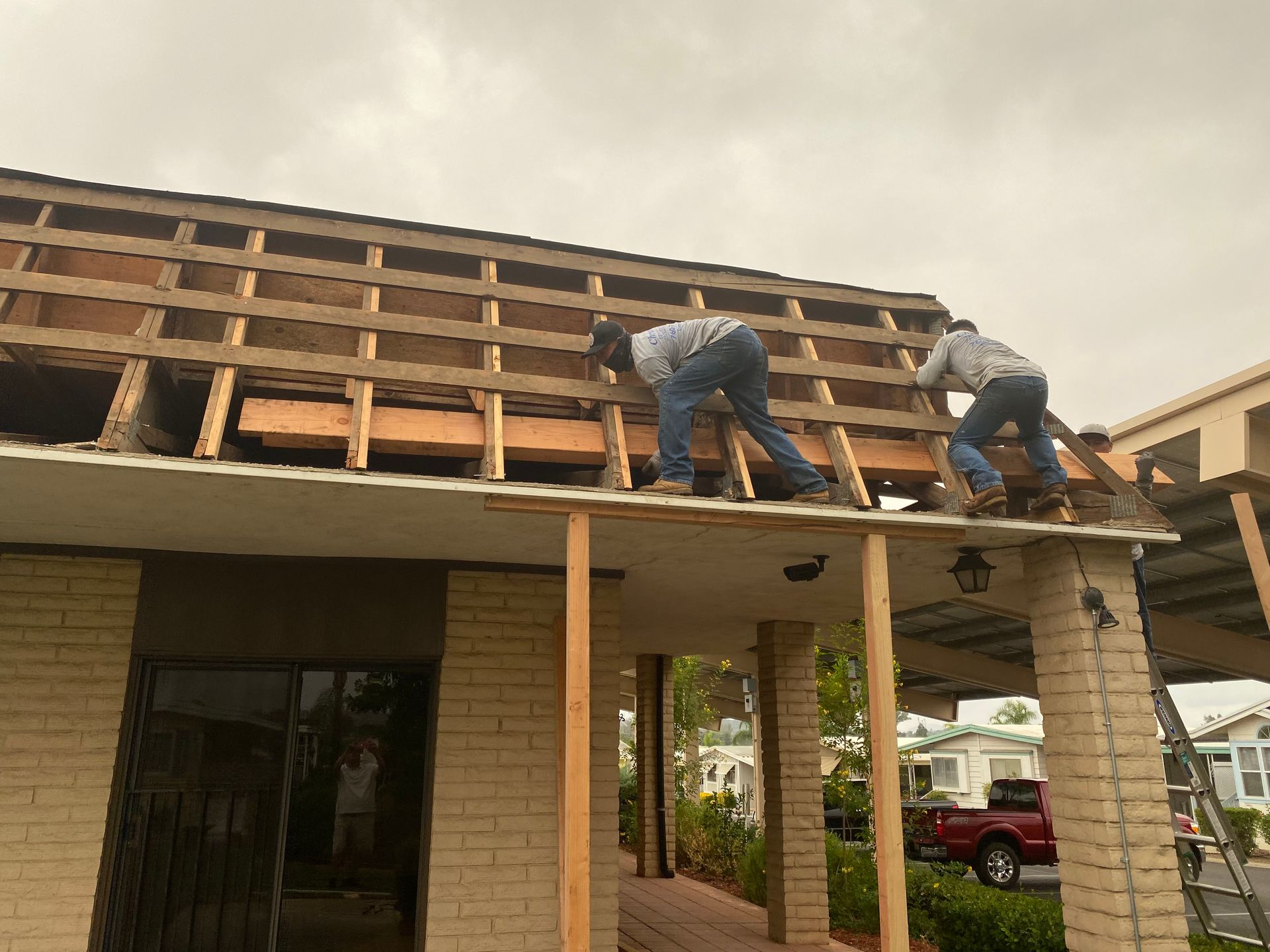 Two construction workers on a roof, removing old material. Beige brick house, cloudy sky.
