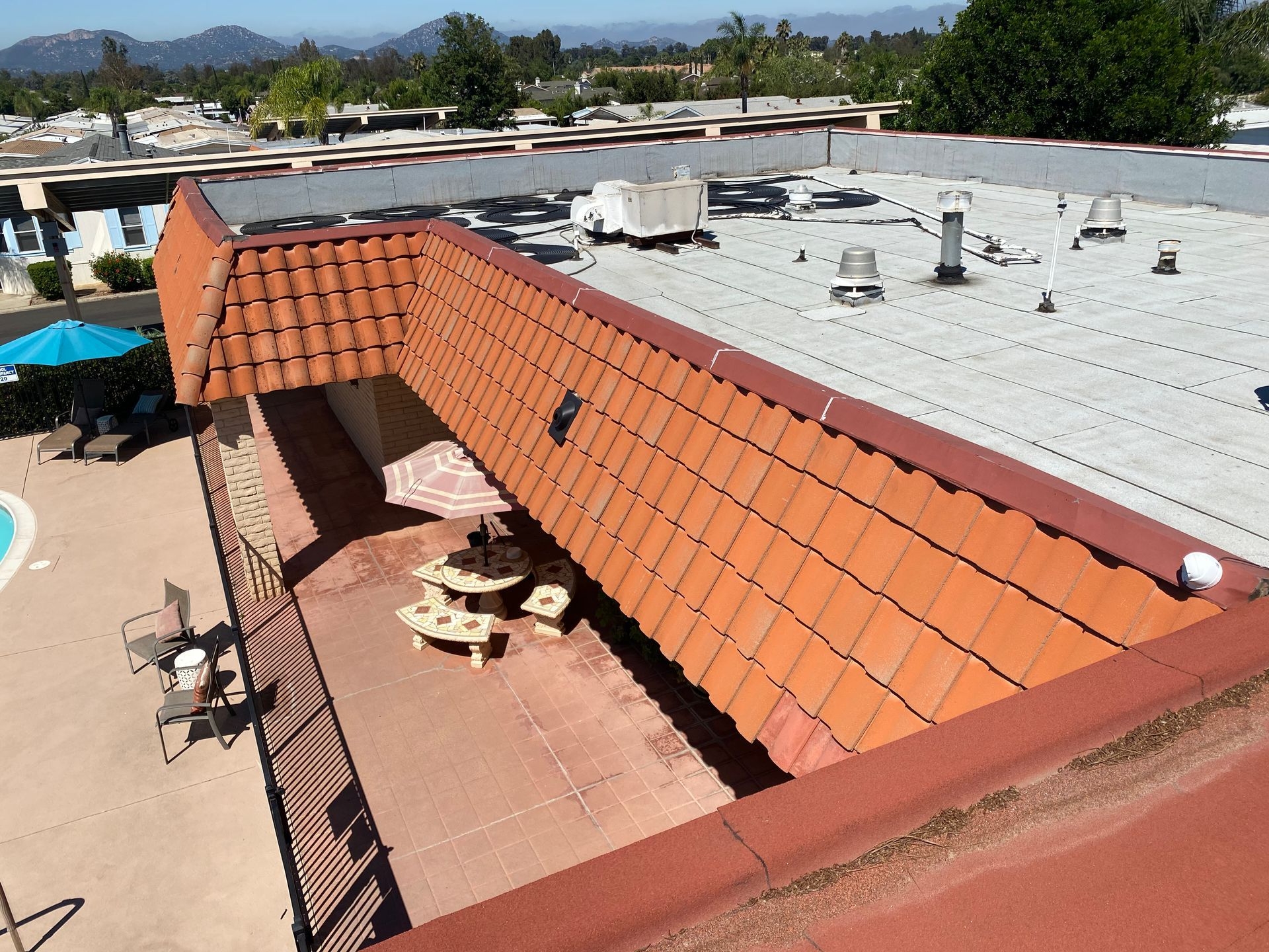 View from rooftop of building with red tile roof section and flat roof. Picnic tables on outdoor deck; pool visible.