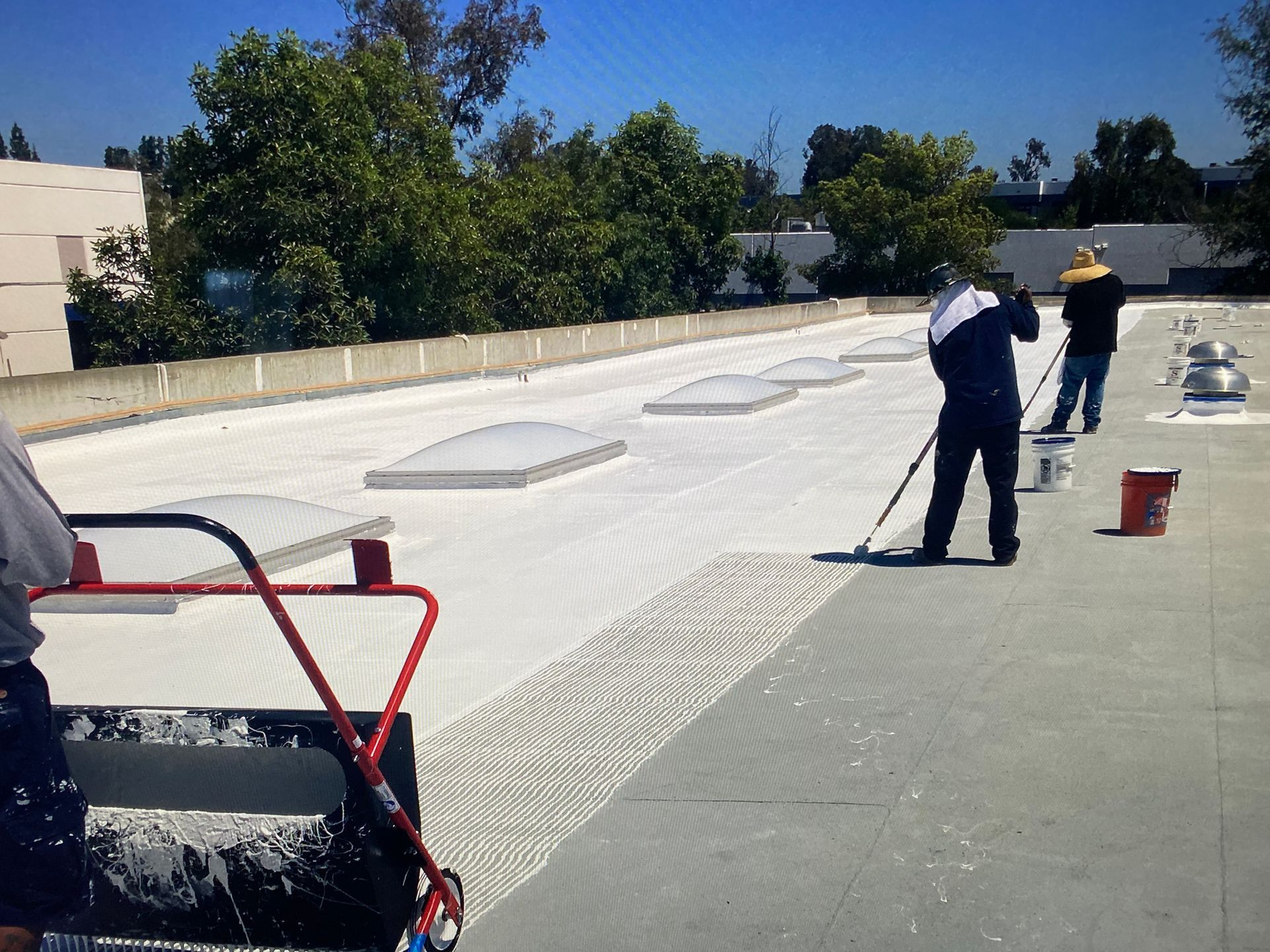 Workers coating a flat roof white. One rolls paint, two others apply with poles. Sunny day.