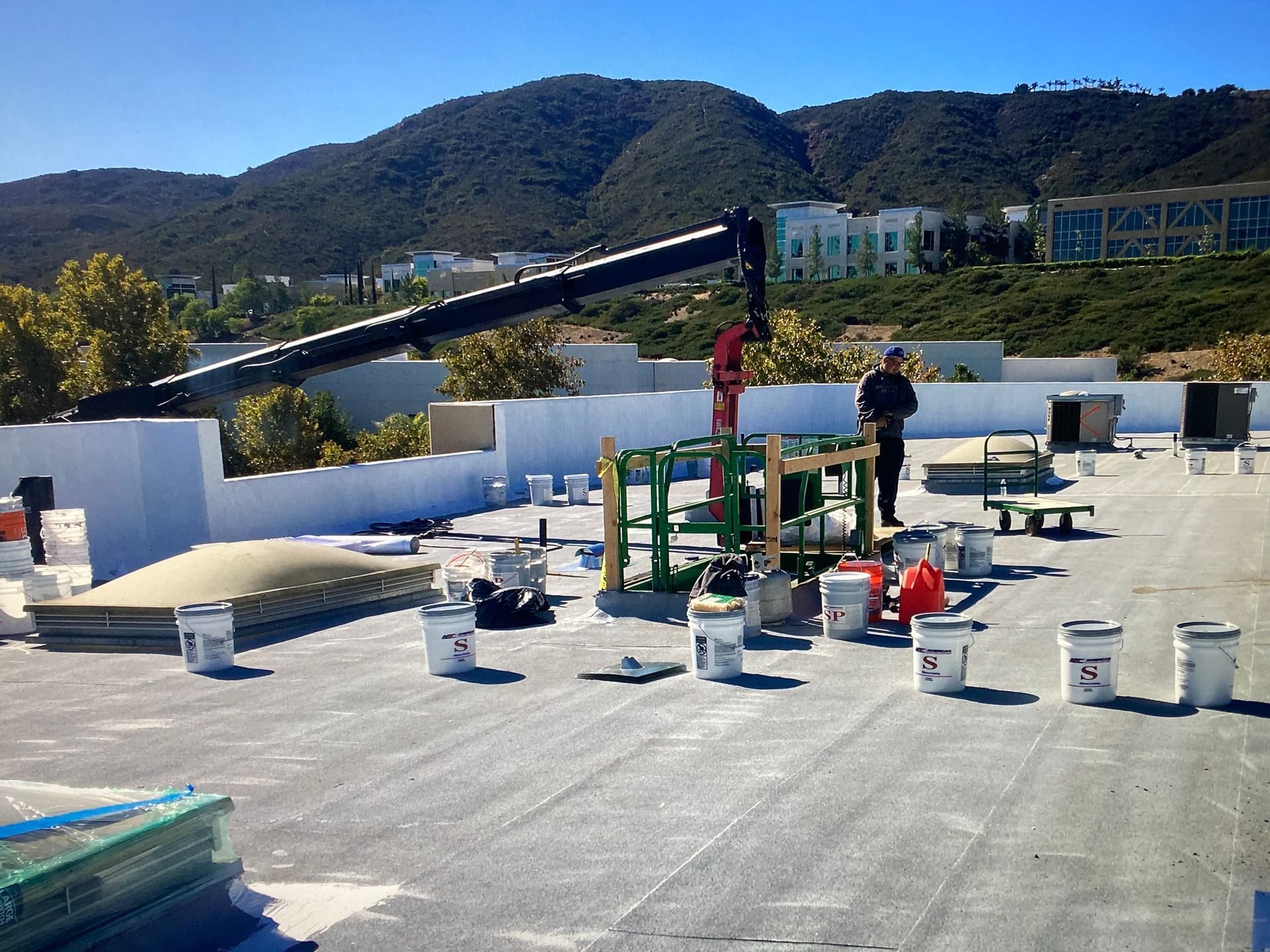 Construction workers on a white roof, using a crane and surrounded by buckets, mountains in the background.