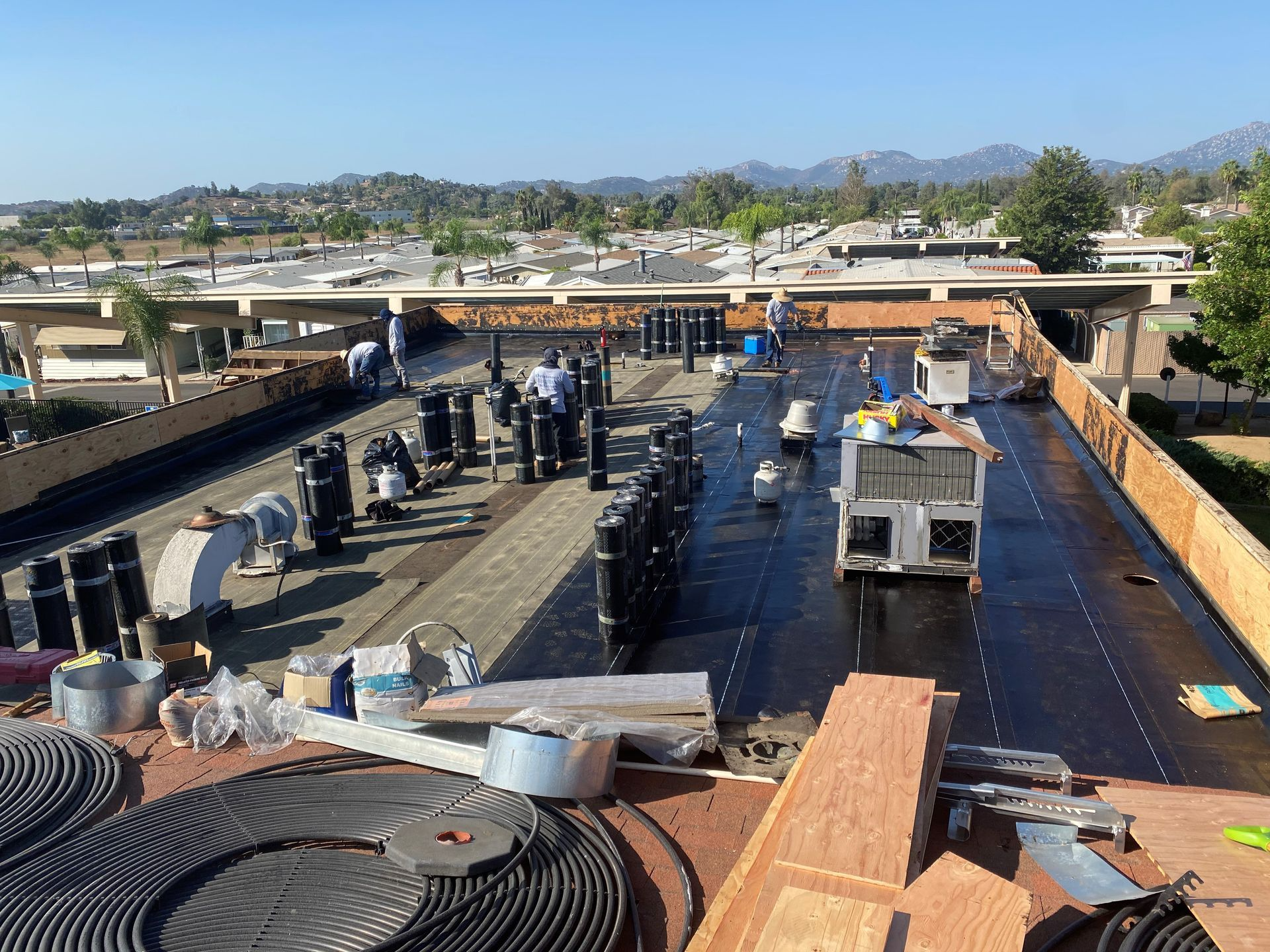 Construction workers on a flat roof, installing roofing materials. Sunny day with distant hills.