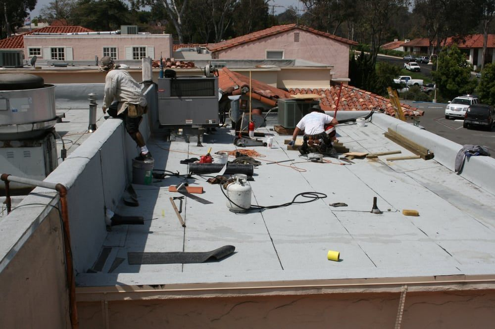 Roofers working on a flat roof, tools and equipment scattered. Buildings and cars in background. Sunny day.