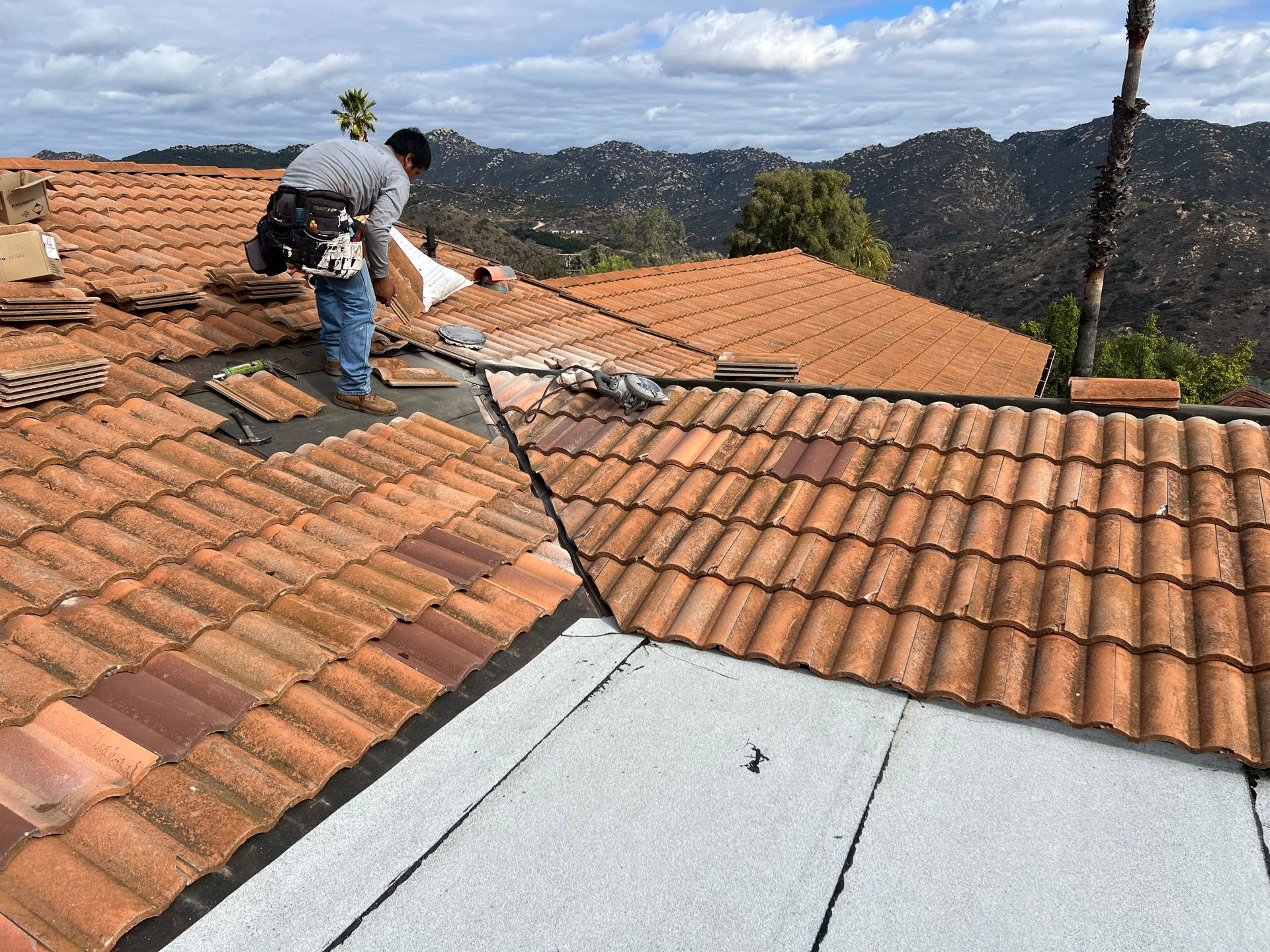 Roofer replacing terracotta tiles on a home roof, mountains in the background on a cloudy day.