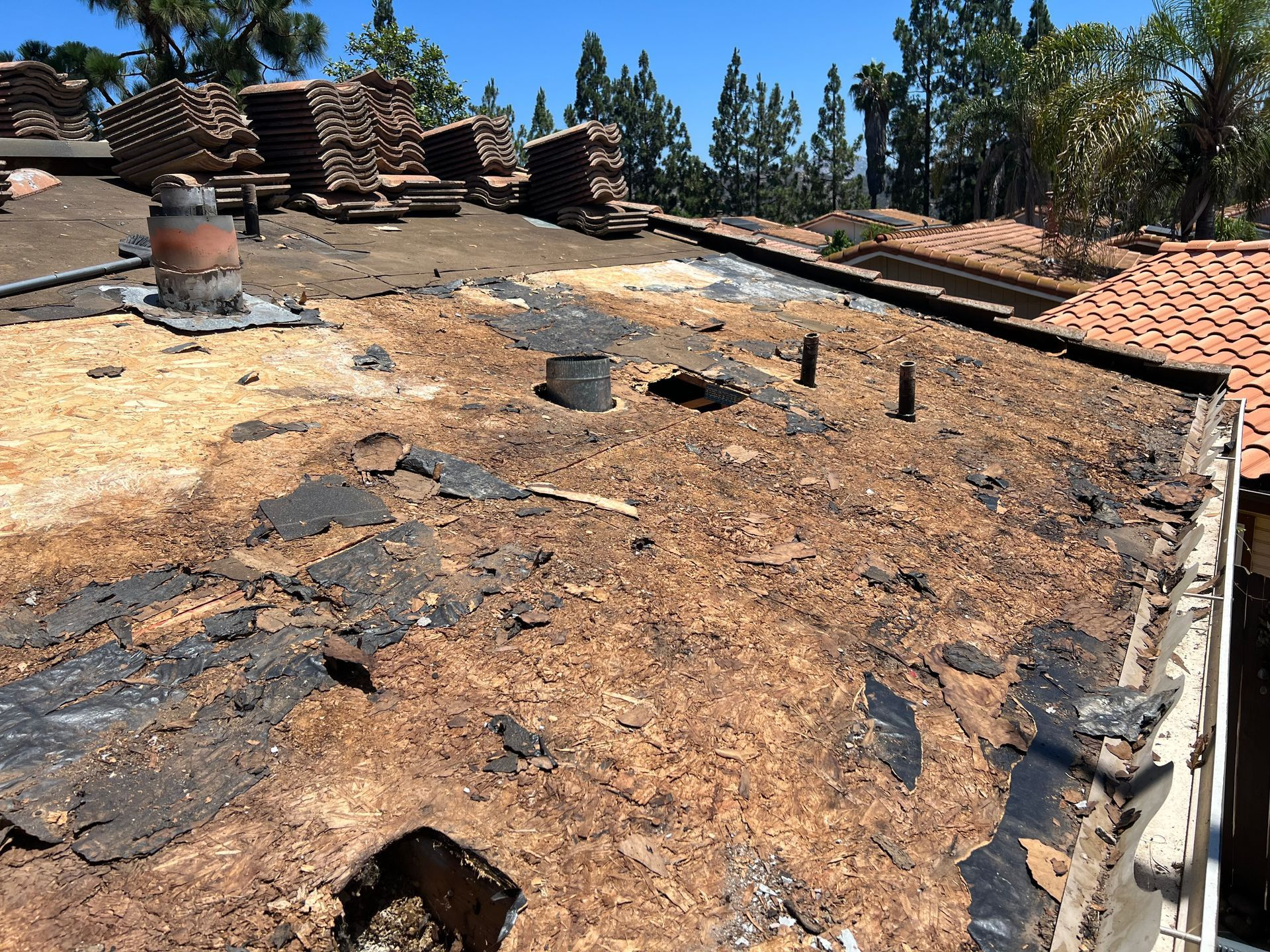 Damaged flat roof with exposed underlayment and stacked tiles, overlooking trees.