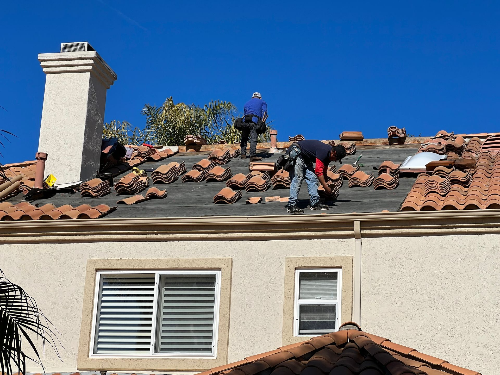 Two roofers replacing clay tiles on a sunny day, visible from a two-story stucco home.