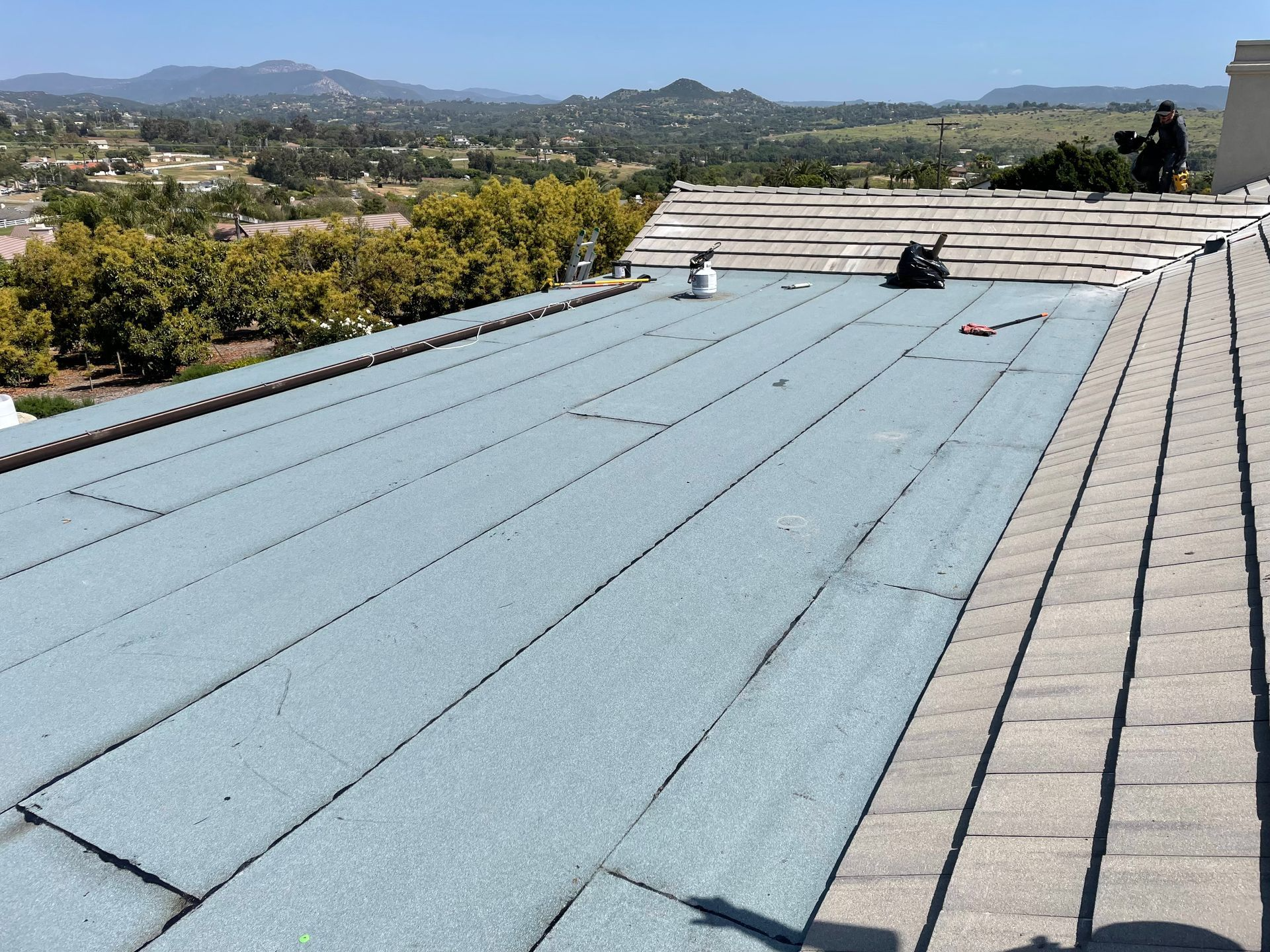 Rooftop with gray shingles and blue, gravel-like material, mountains in background.
