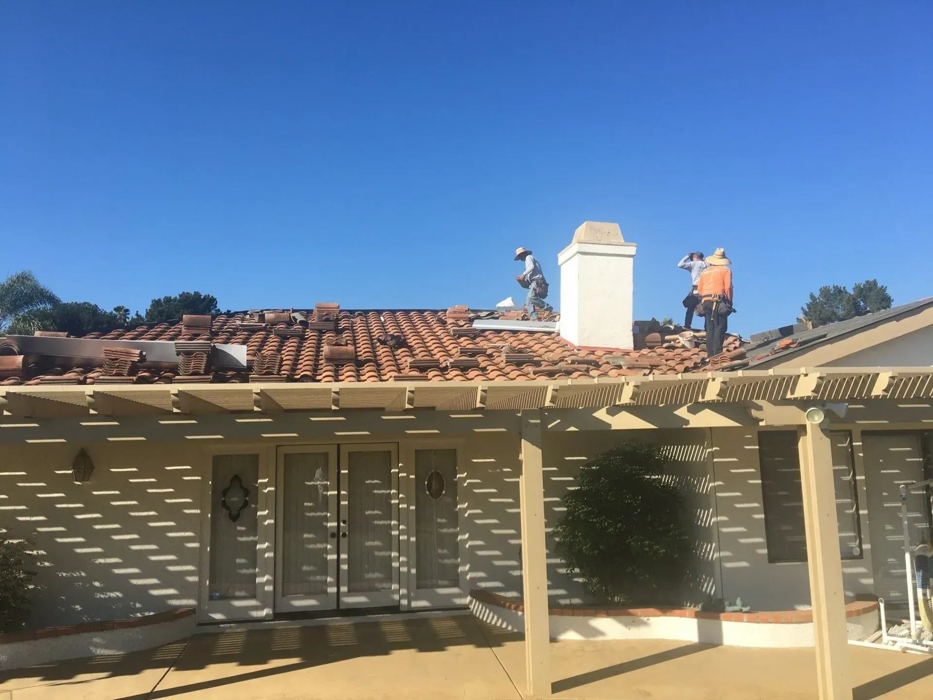 Workers on a roof repairing tiles; chimney, pergola, and a clear blue sky.