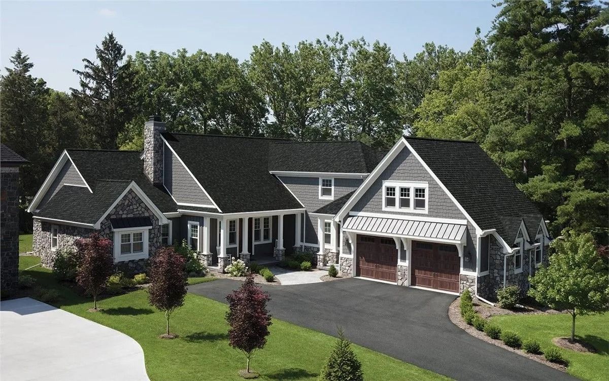 Gray, multi-gabled house with stone accents, a long driveway, and lush green landscaping.
