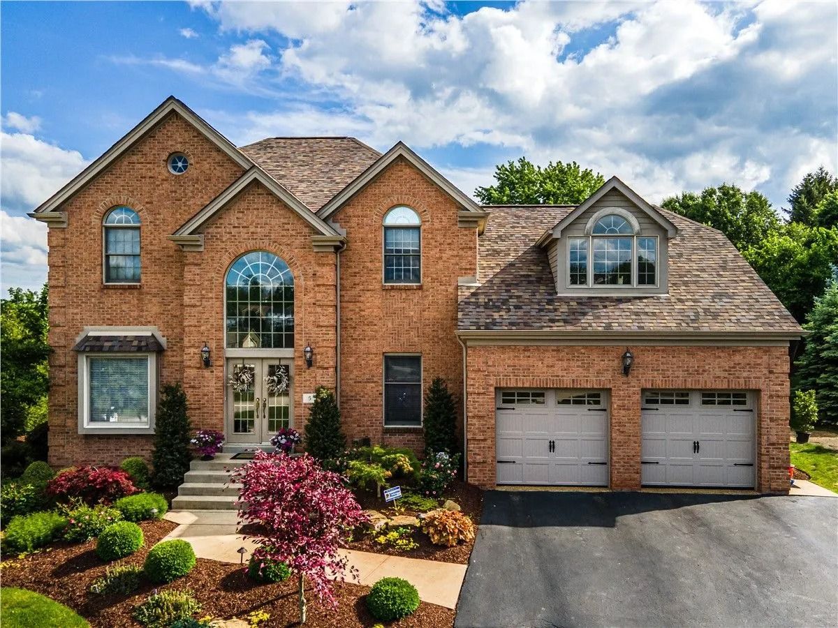 Two-story brick house with a two-car garage, arched windows, and lush landscaping under a cloudy sky.