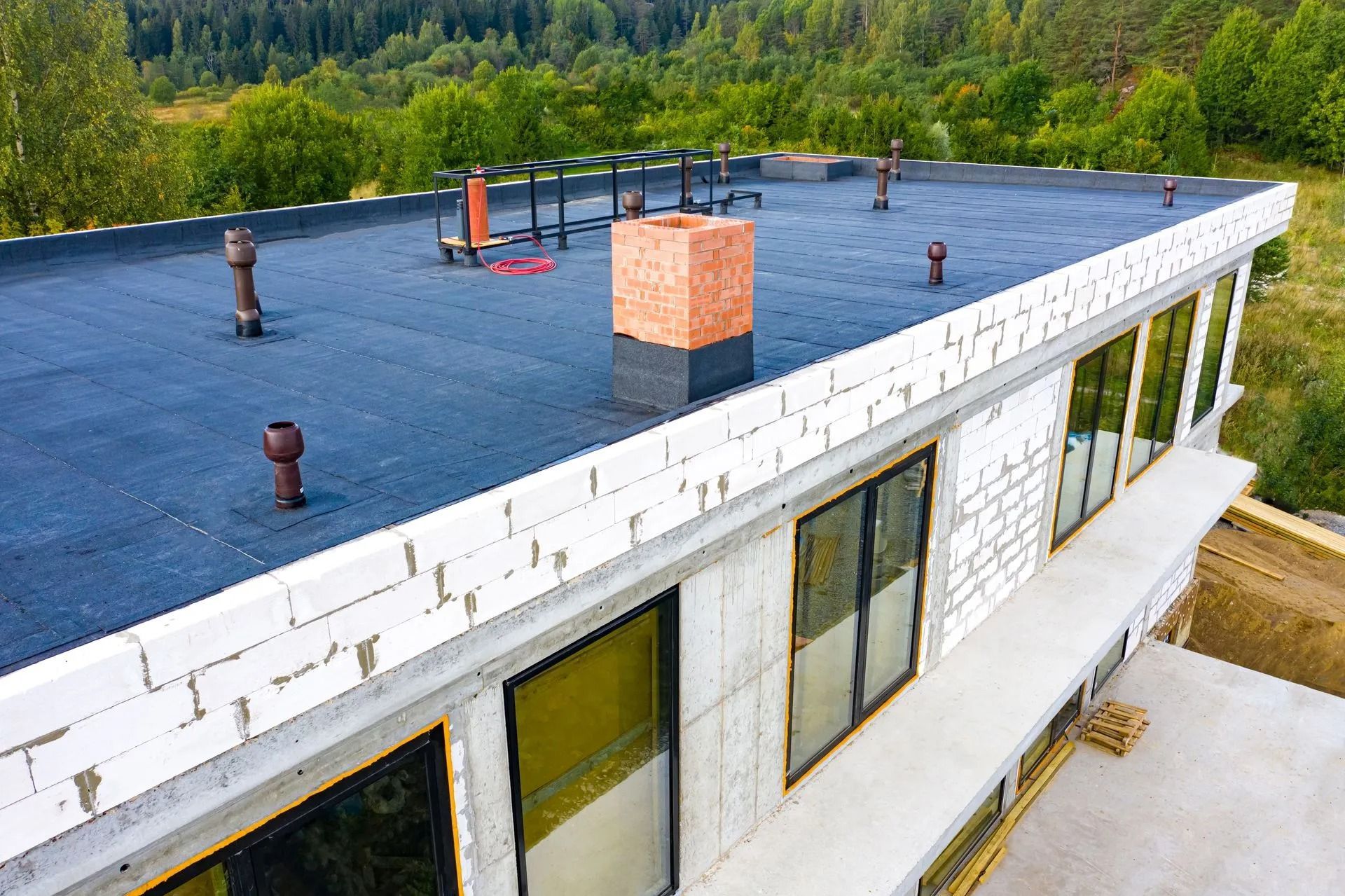 Modern building under construction with a flat black roof, brick chimney, and large windows, surrounded by trees.