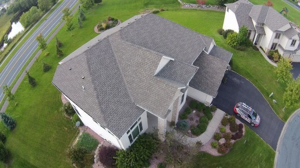 Aerial view of a light-colored house with gray roof, driveway, and a car. Lush green lawn and trees surround it.